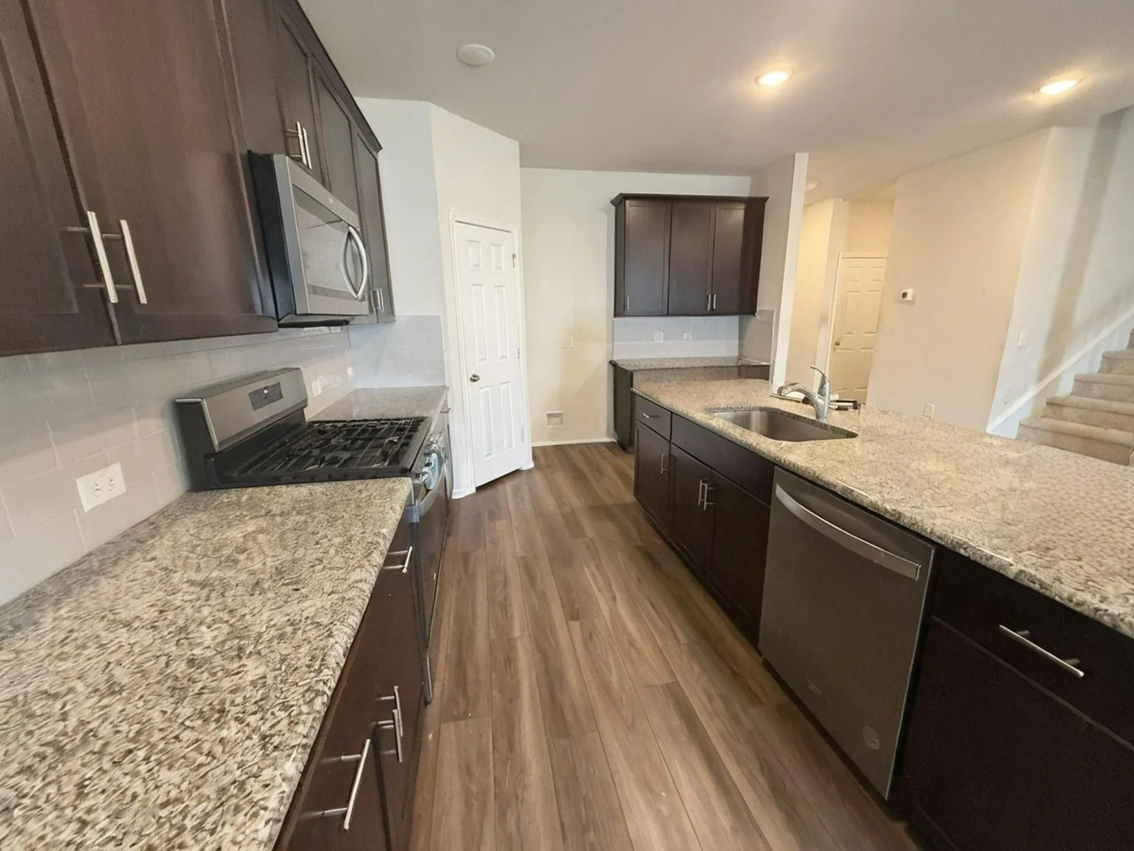 Galley kitchen featuring wood-finish flooring, dark wood cabinetry, light-toned countertops, stainless steel appliances, and a tile backsplash