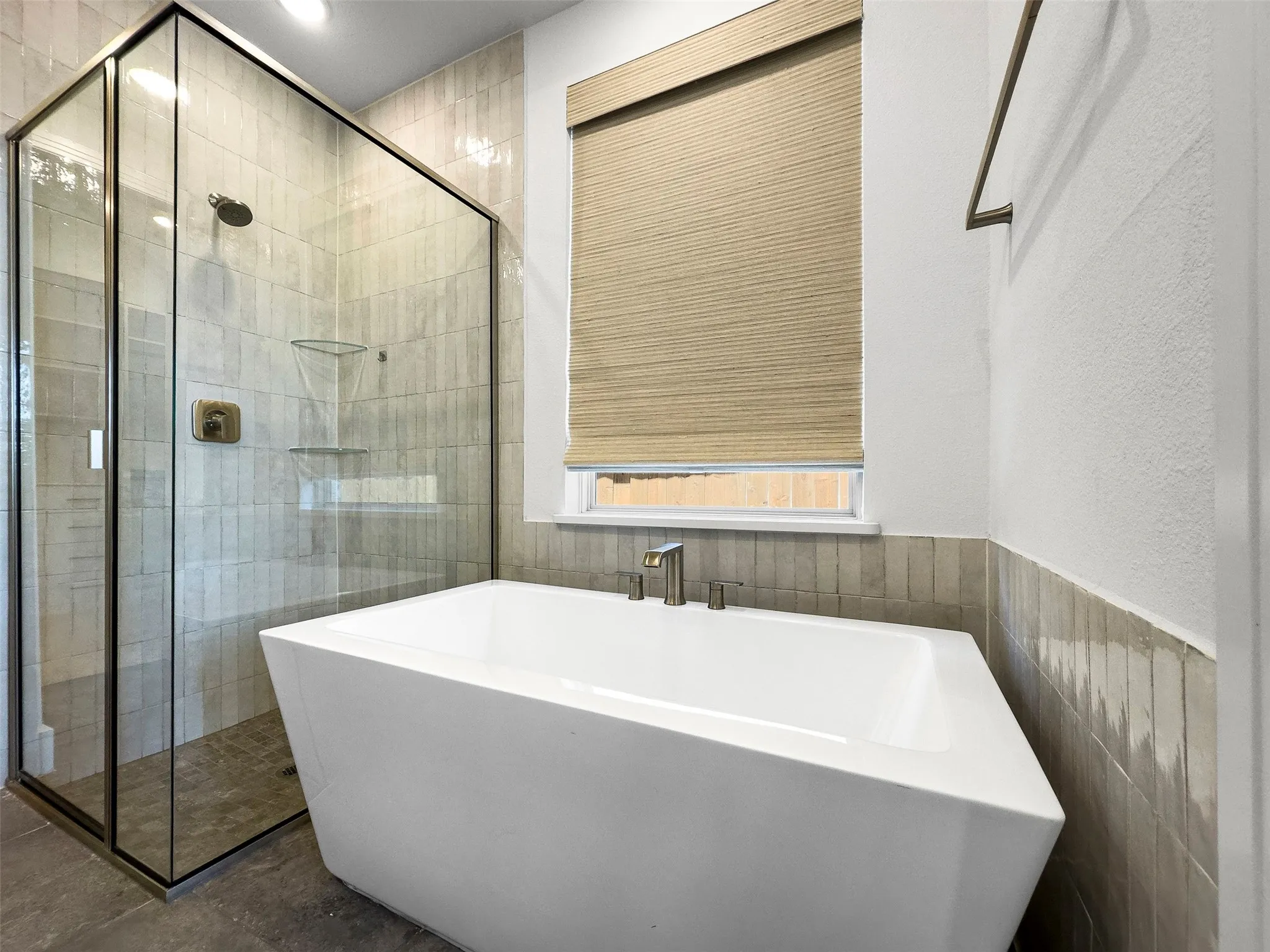 Freestanding soaking tub with brushed nickel fixtures, glass-enclosed shower with tiled walls and bronze-finish hardware, and a window with a woven Roman shade