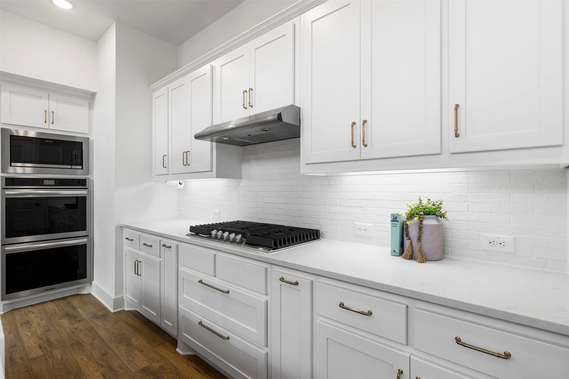 White shaker cabinetry, built-in double ovens, stainless steel appliances, gas cooktop, and wood-finish flooring