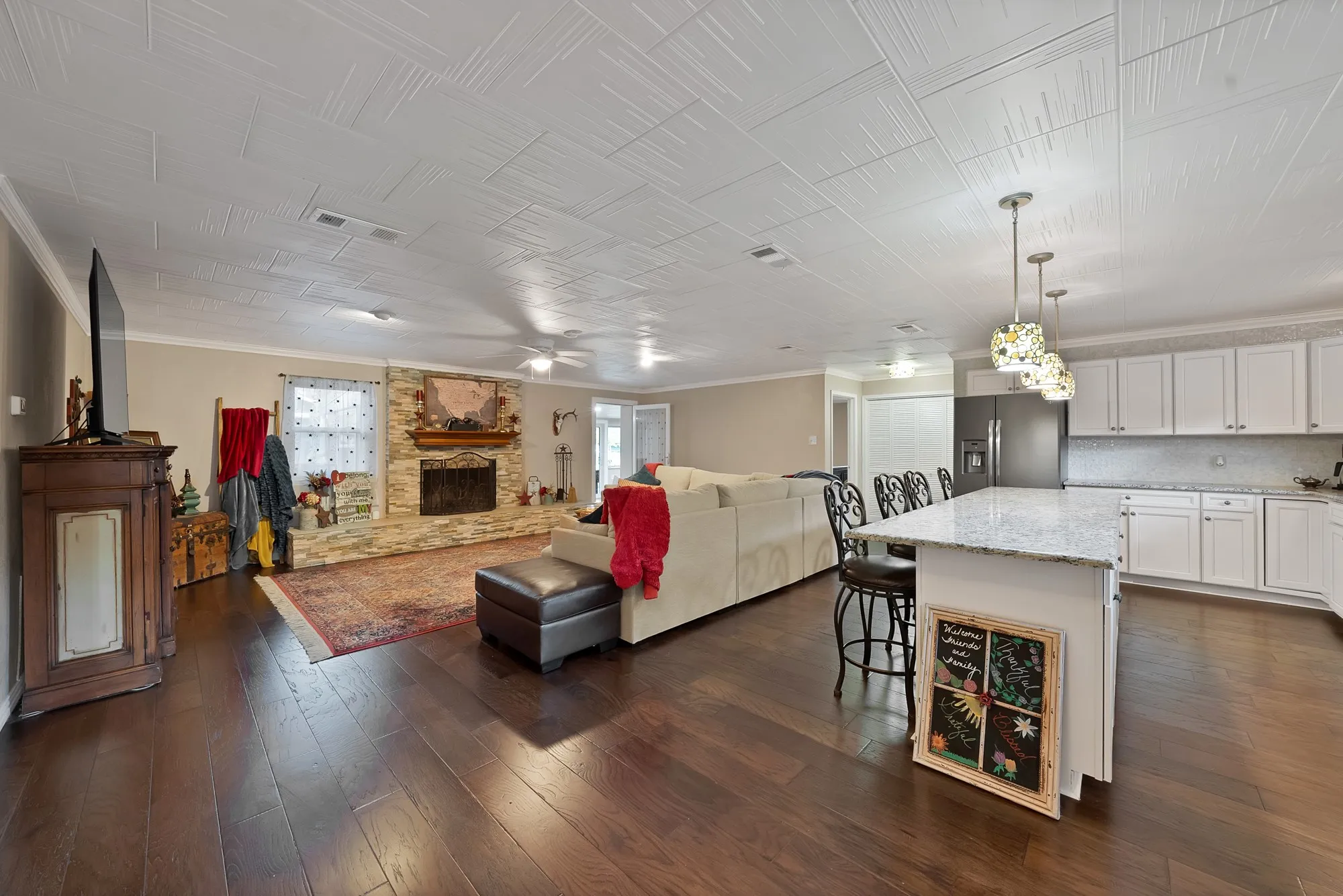 Open-concept interior featuring wood-finish flooring, a stone-clad fireplace, a kitchen island with seating, pendant lighting, and white cabinetry