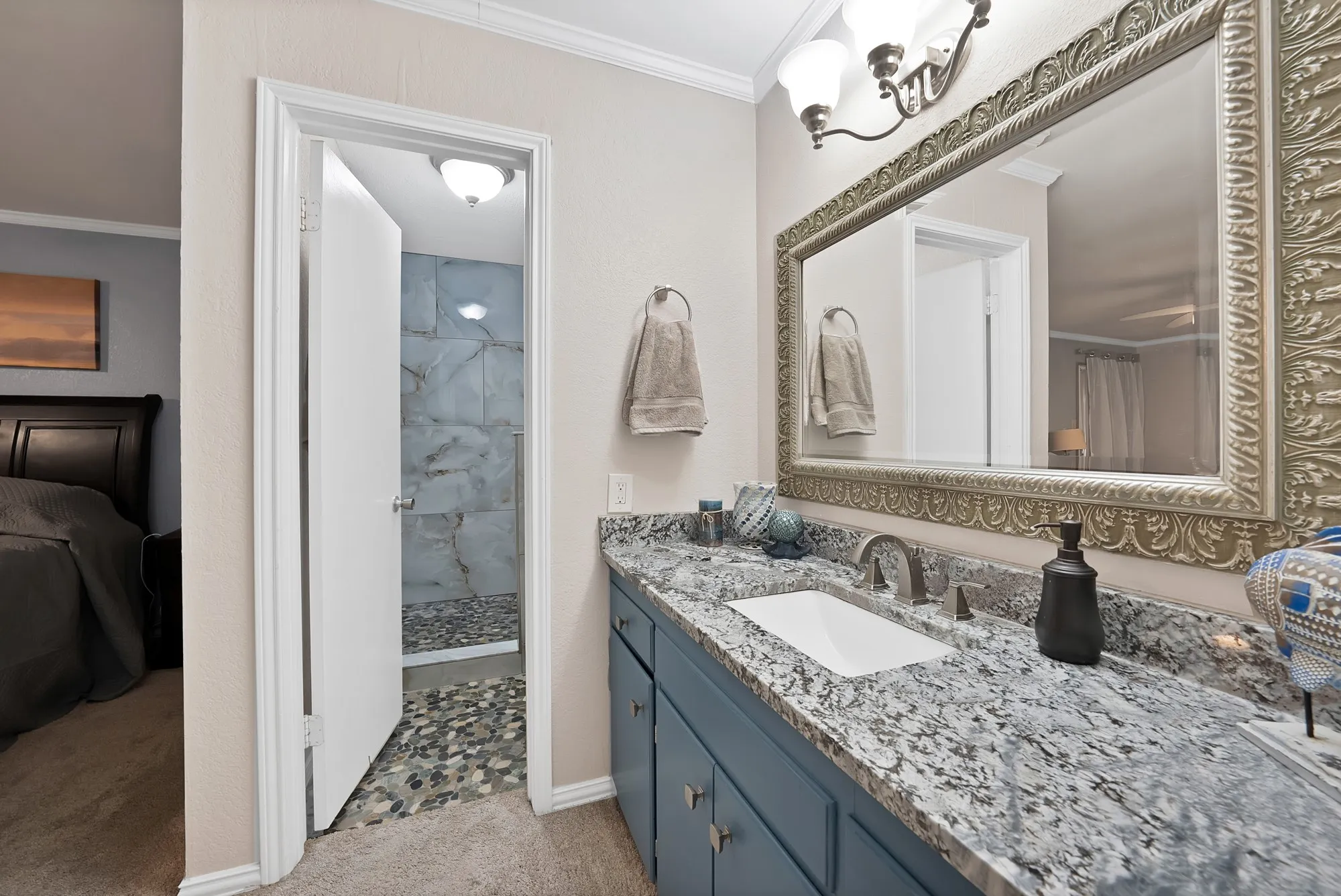 Vanity with light-toned countertop and integrated sink, decorative framed mirror, blue base cabinetry, and a walk-in shower with stone tile flooring and large format wall tiles
