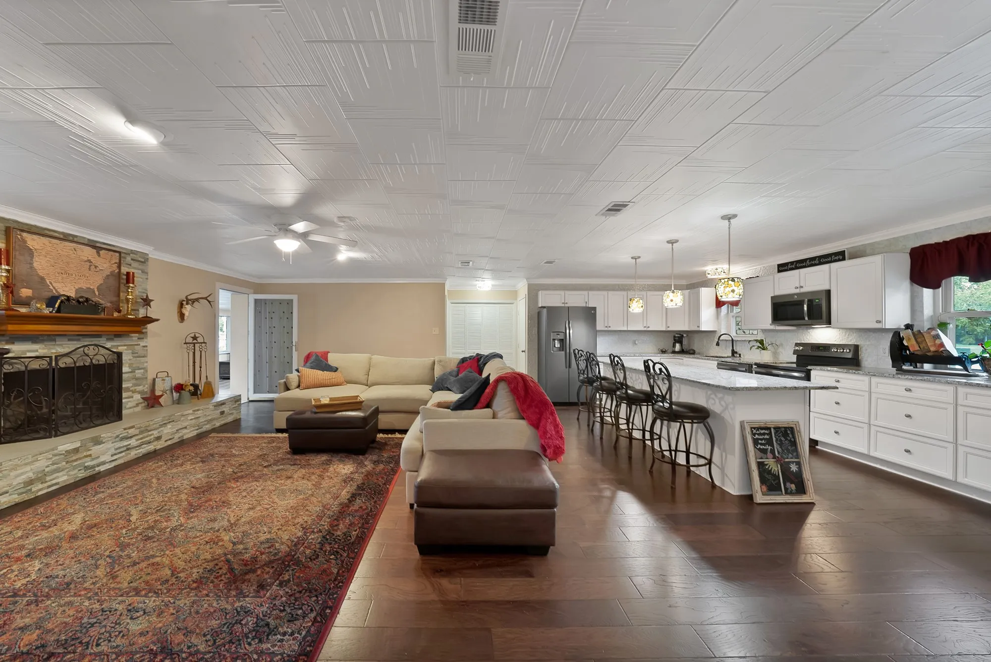 Open-concept living space featuring wood-finish flooring, a stone-faced fireplace with a wood mantel, and a kitchen with white cabinetry and an island
