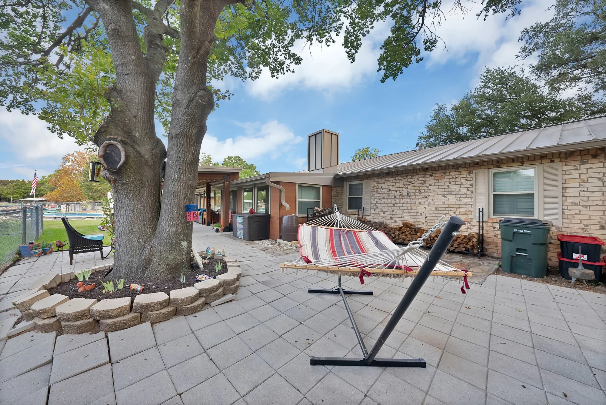 Expansive paved patio area featuring a large mature tree with stone-bordered planting, a brick-clad exterior, and a standing seam metal roof