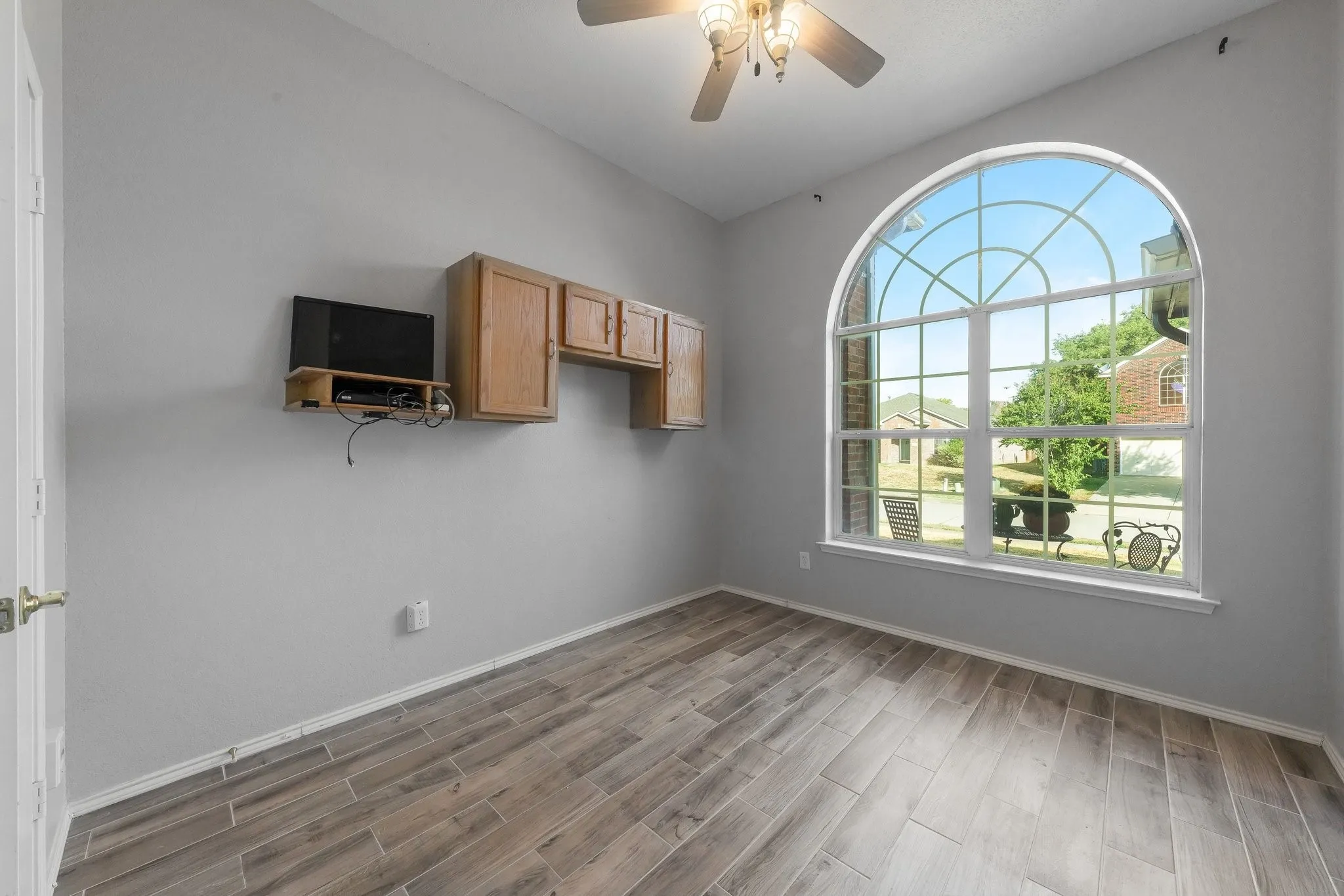 Arch-top window with decorative grid, light wood-finish flooring, ceiling fan with integrated lighting, and built-in wood cabinetry