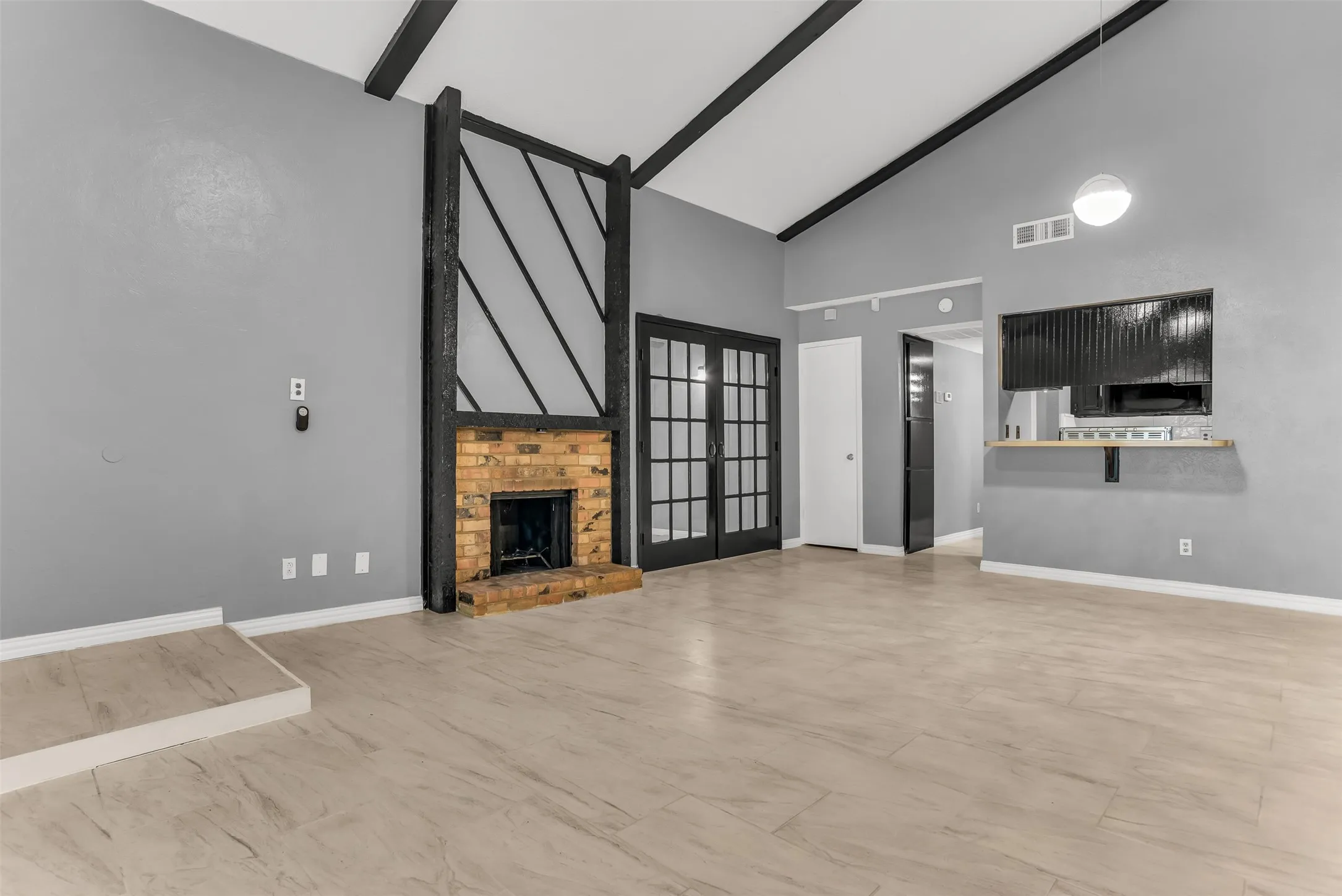 Vaulted ceilings with exposed beams, brick fireplace with decorative wood surround, wood-finish flooring, and black French doors with paneled glass