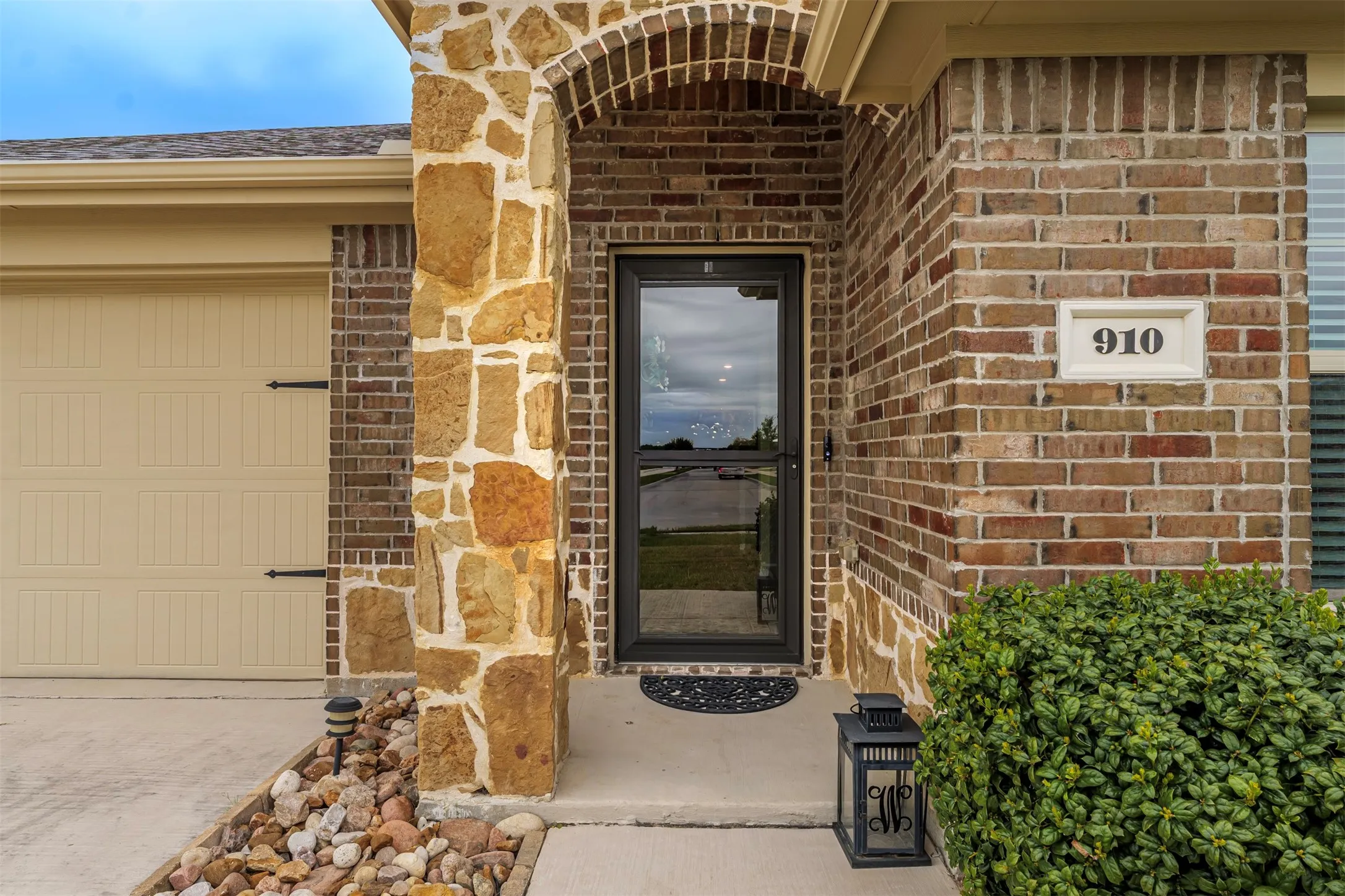 Brick exterior with a stone accent column, arched entryway, garage door with decorative hardware, and a glass-paneled entry door