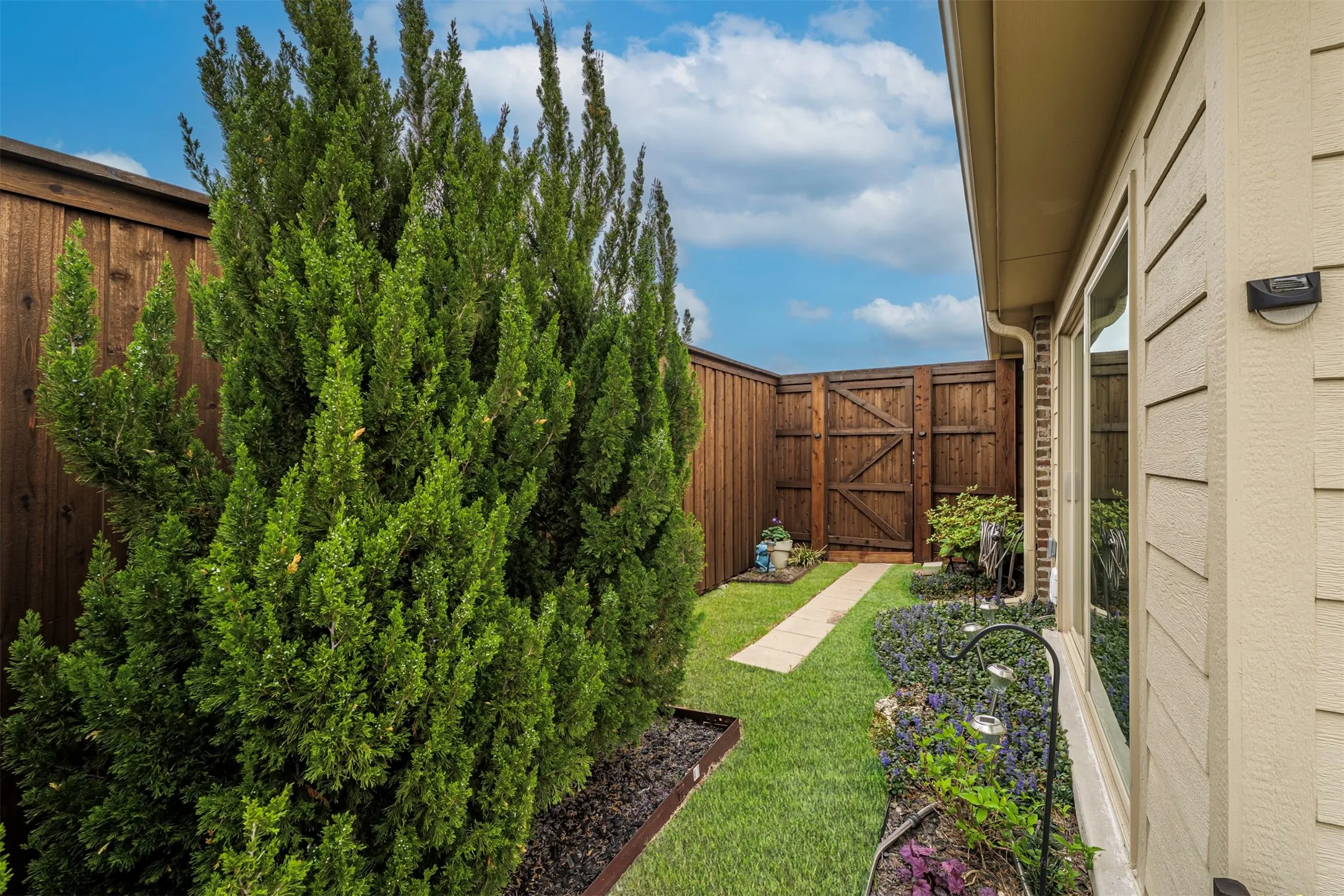 Landscaped side yard featuring a privacy fence, established shrubbery, a grass pathway, and a side gate