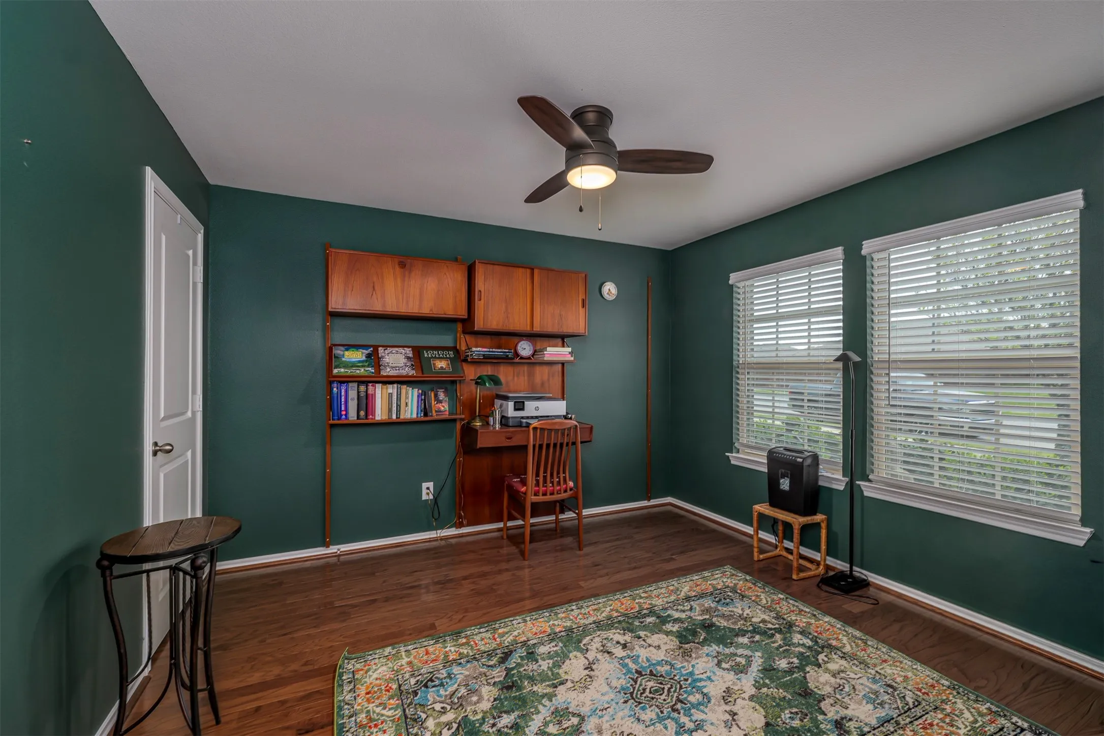 Hardwood flooring, built-in shelving, two windows with blinds, ceiling fan with integrated lighting, and a white paneled door