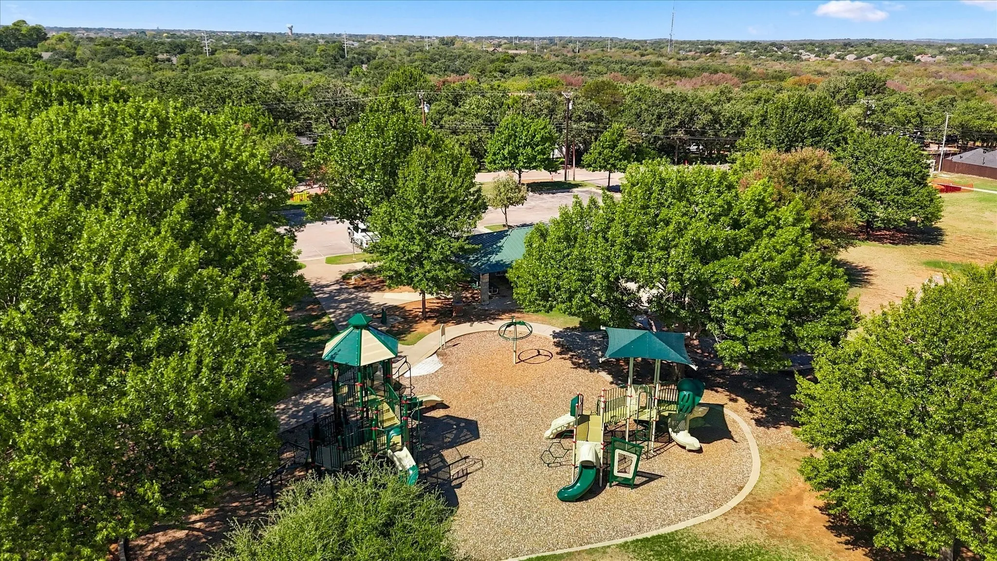 Playground area featuring multiple slides, climbing structures, and a covered seating area, surrounded by mature trees
