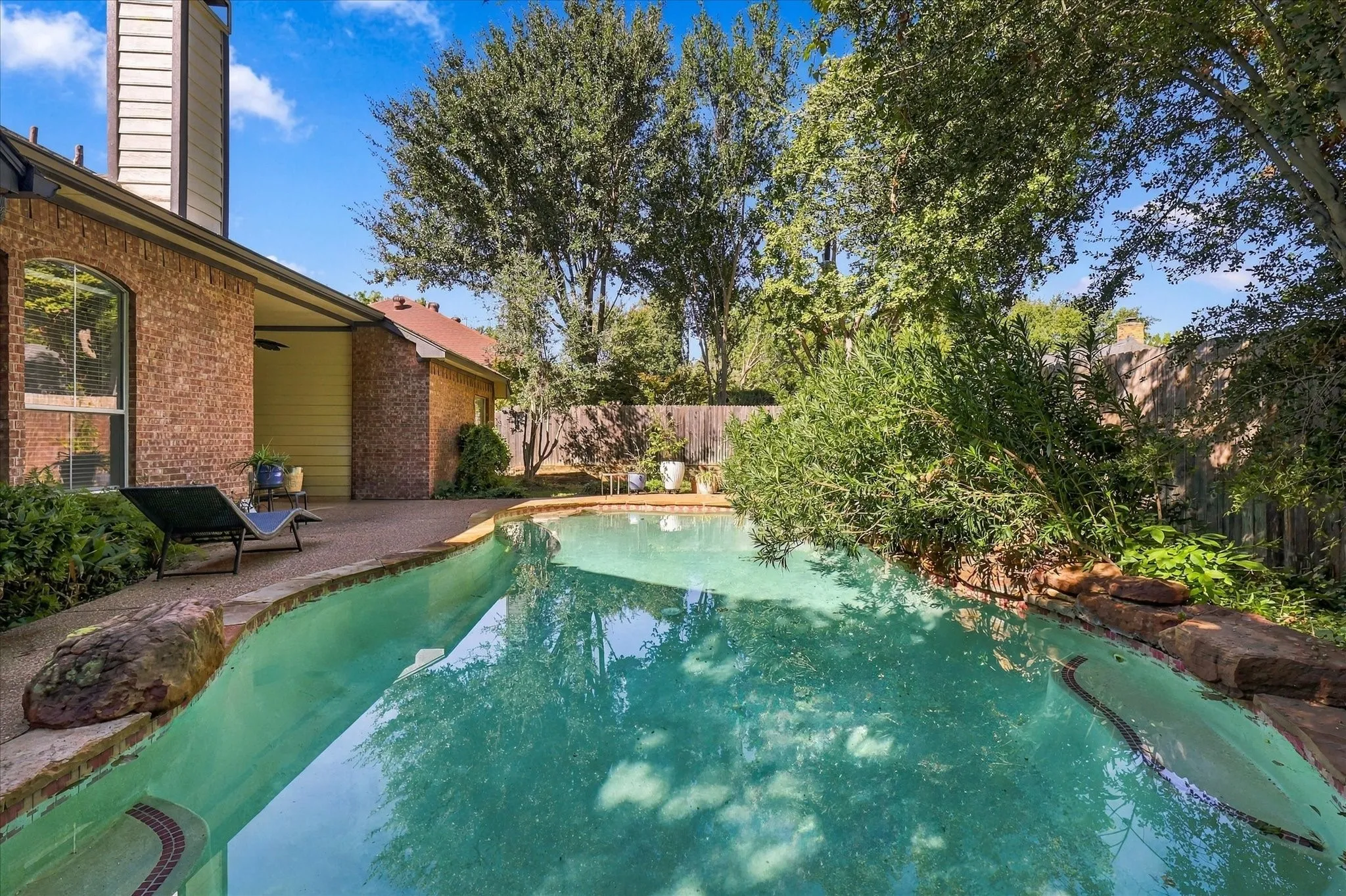 Brick exterior with a covered patio, complemented by a swimming pool with stone coping and lush perimeter landscaping