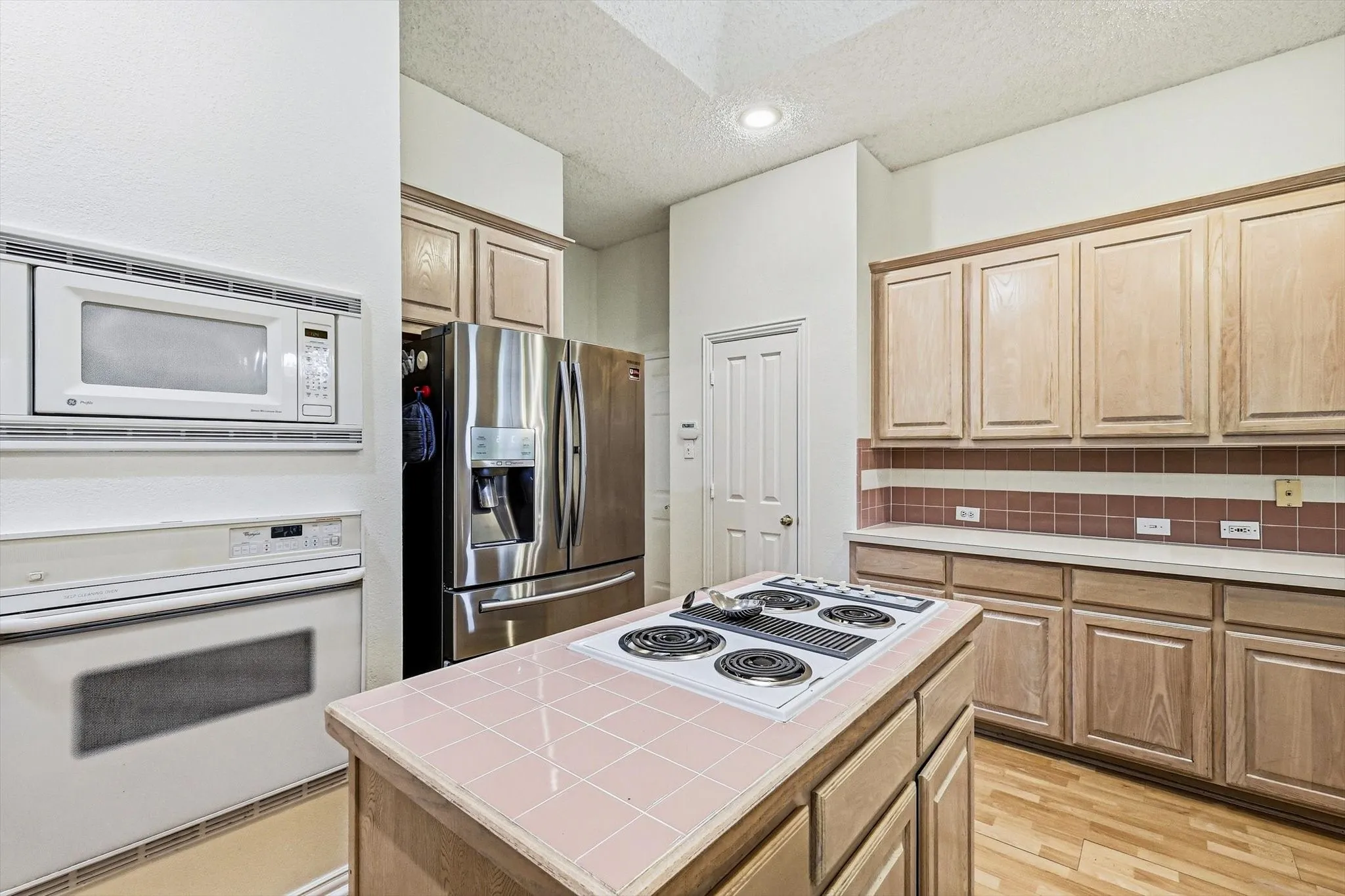 Kitchen featuring light wood cabinetry, a tiled center island, stainless steel refrigerator, built-in microwave, and wood-finish flooring