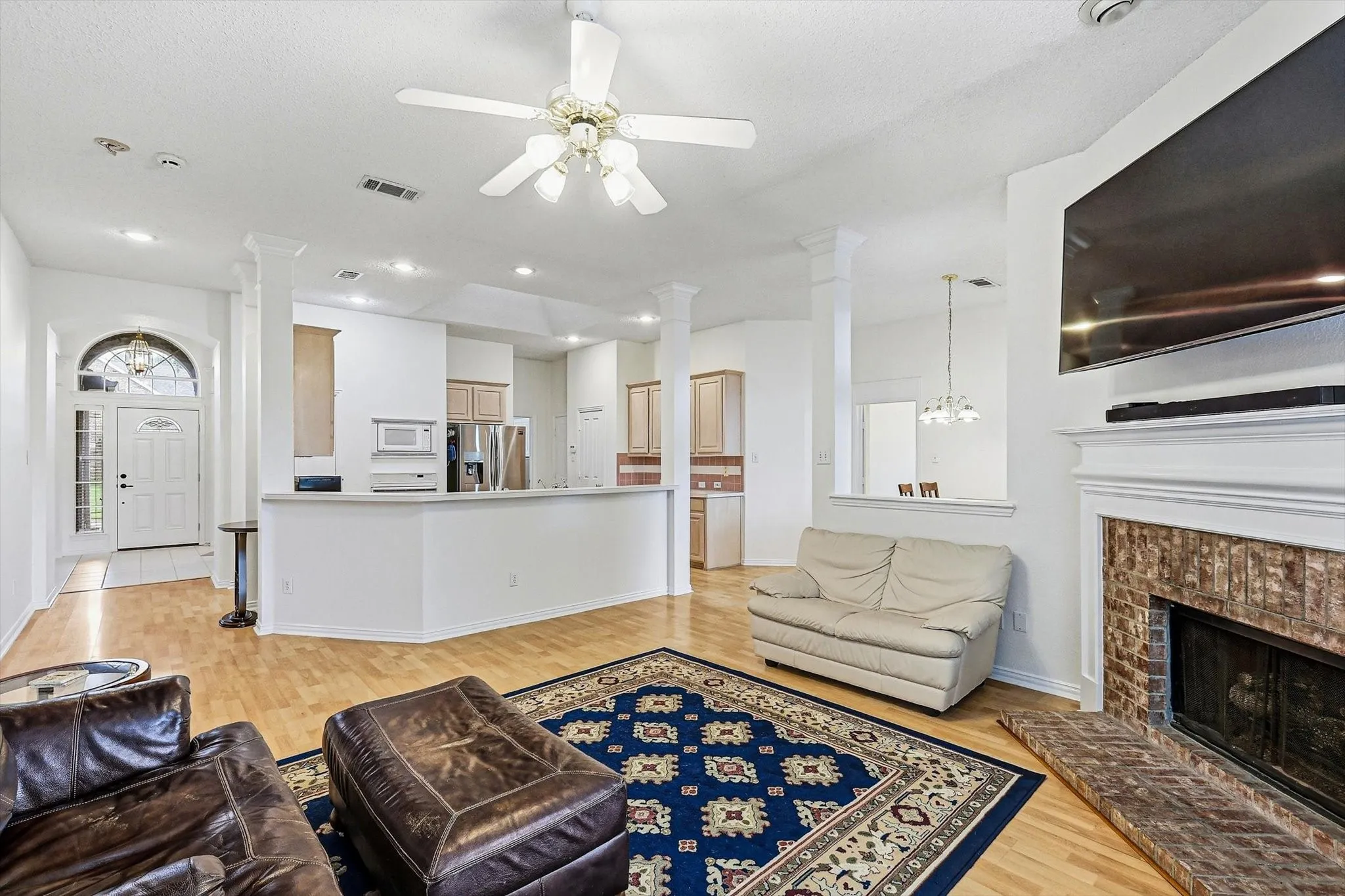 Open-concept living area featuring wood-finish flooring, a brick fireplace with a white mantel, and a kitchen with light wood cabinetry and stainless steel appliances