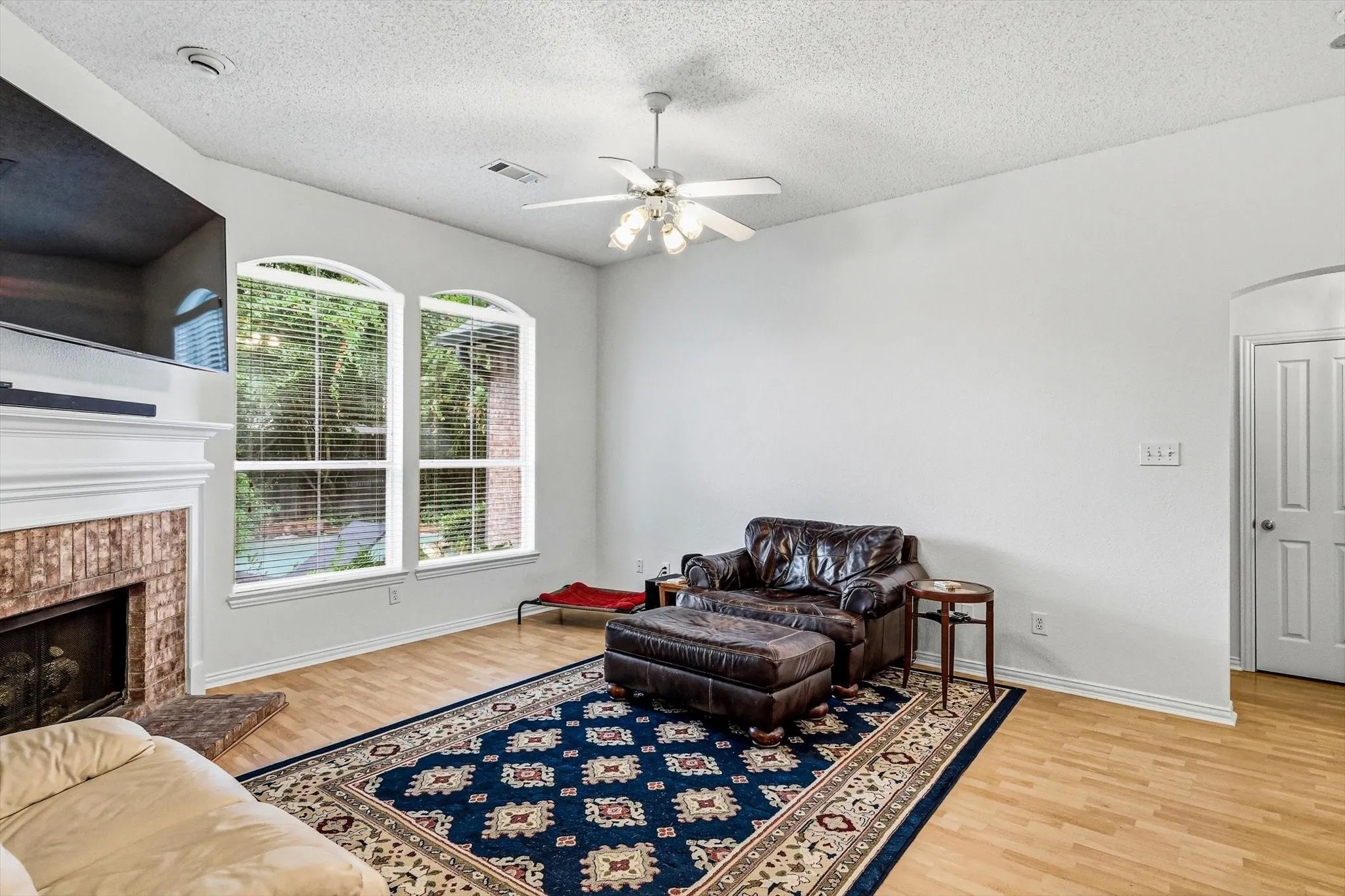 Hardwood-finish flooring, brick-surround fireplace with a white mantel, two arched windows, ceiling fan, and white paneled door