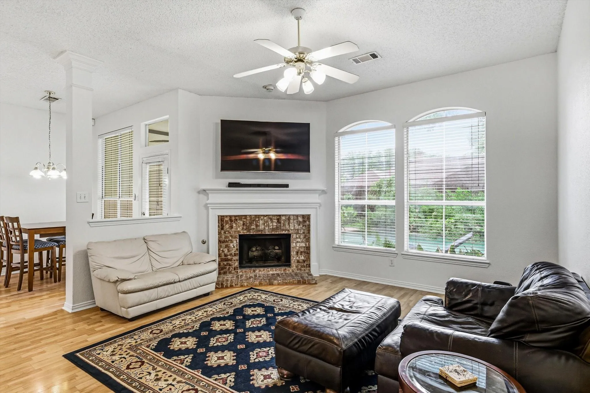 Living room featuring hardwood-finish flooring, a ceiling fan, a fireplace with a brick surround, and arched windows