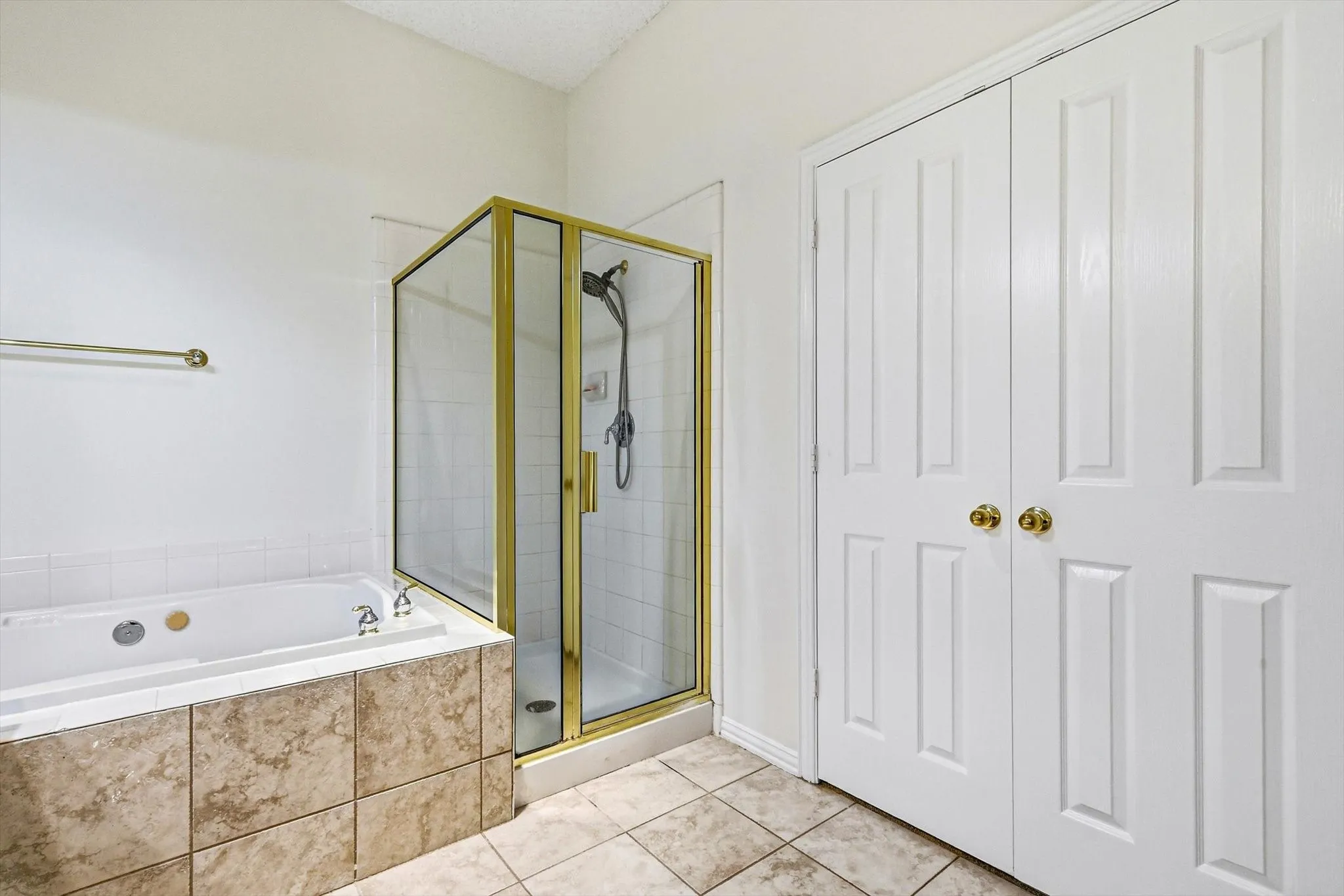 Jetted soaking tub with tiled surround, glass-enclosed shower with gold-finish frame, and paneled bifold doors