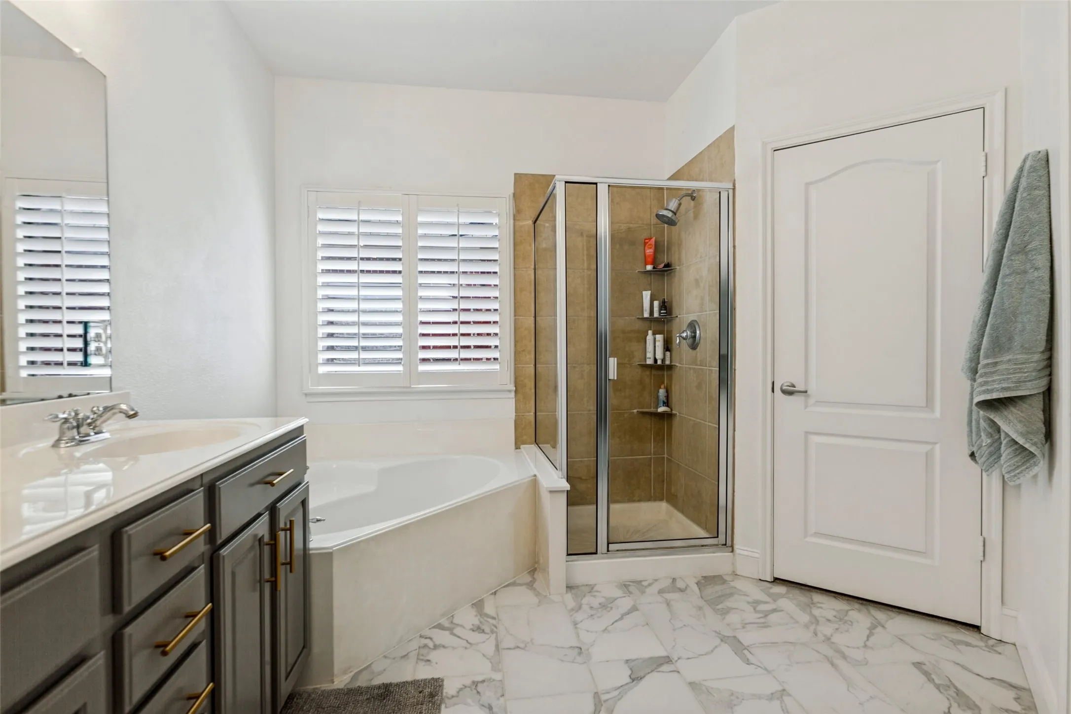 New White marble-look flooring, built-in jetted soaking tub, glass-enclosed shower with tile surround, vanity with dark cabinetry, and interior plantation shutters