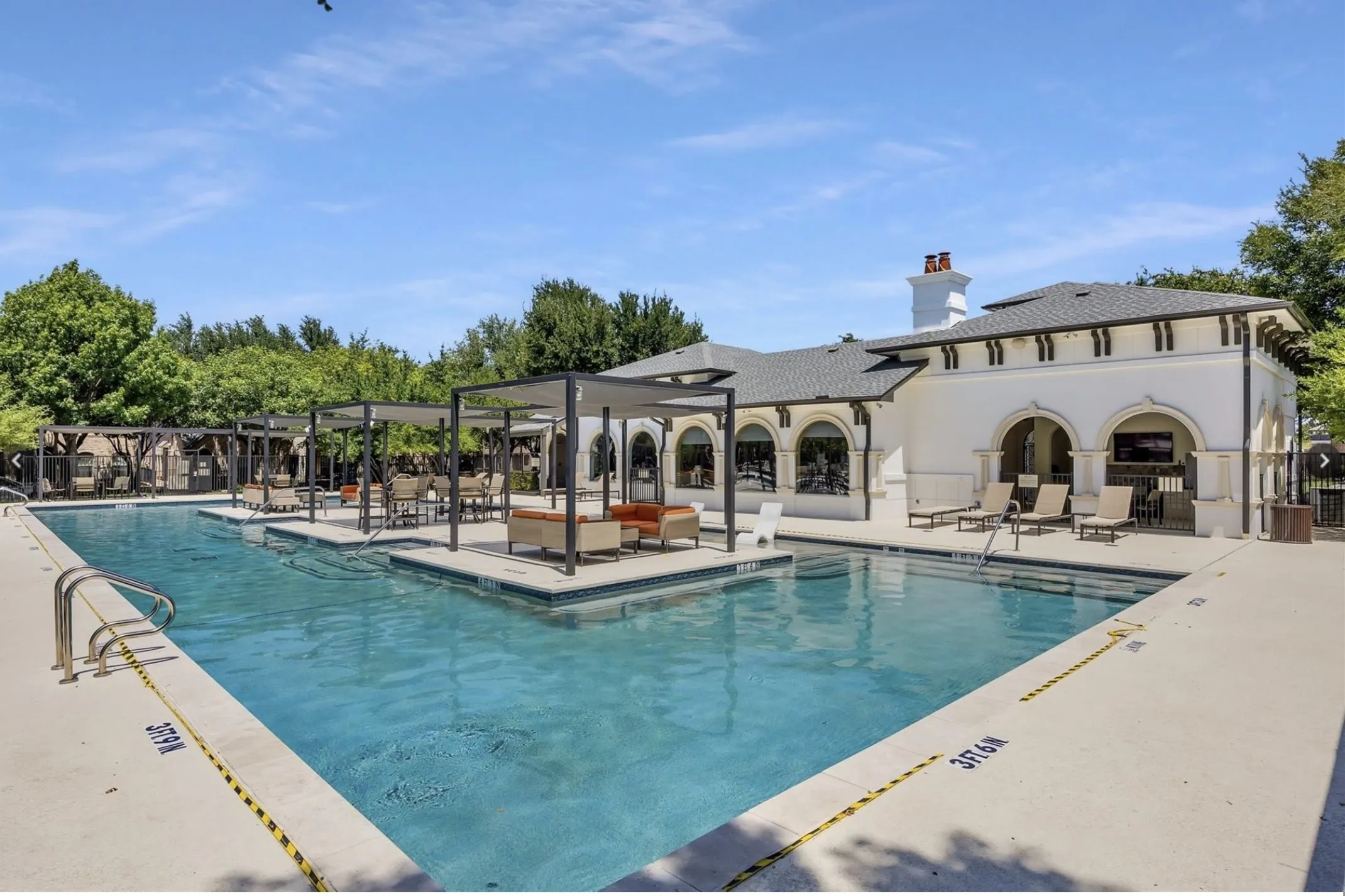 Expansive swimming pool with integrated tanning ledges, multiple shade pergolas, and a stucco-finished building featuring arched openings and a dark tile roof