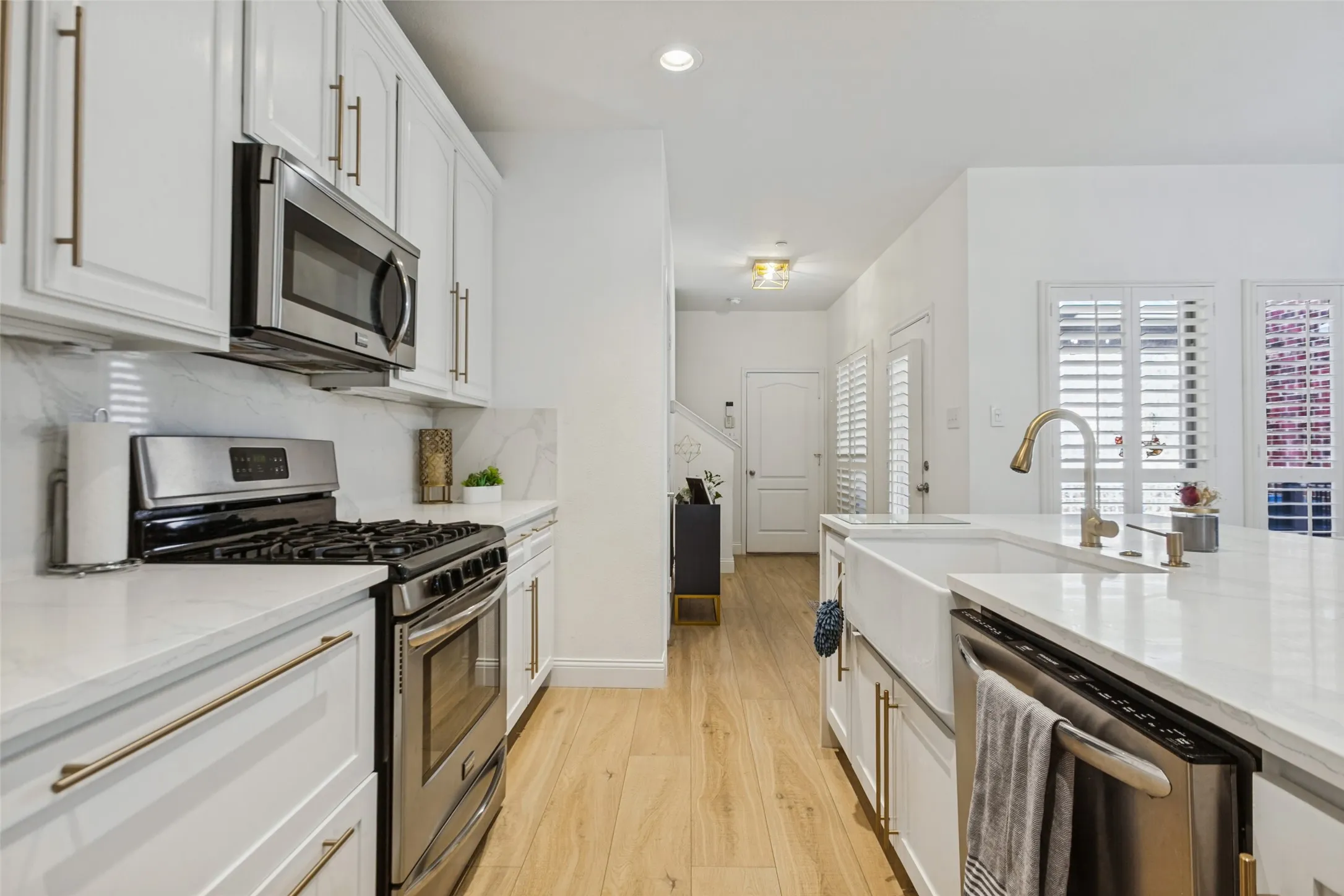 Custom white shaker cabinetry with gold-tone hardware, stainless steel appliances, white countertop surfaces, wood-finish flooring, and a farmhouse-style sink with a gold-tone faucet