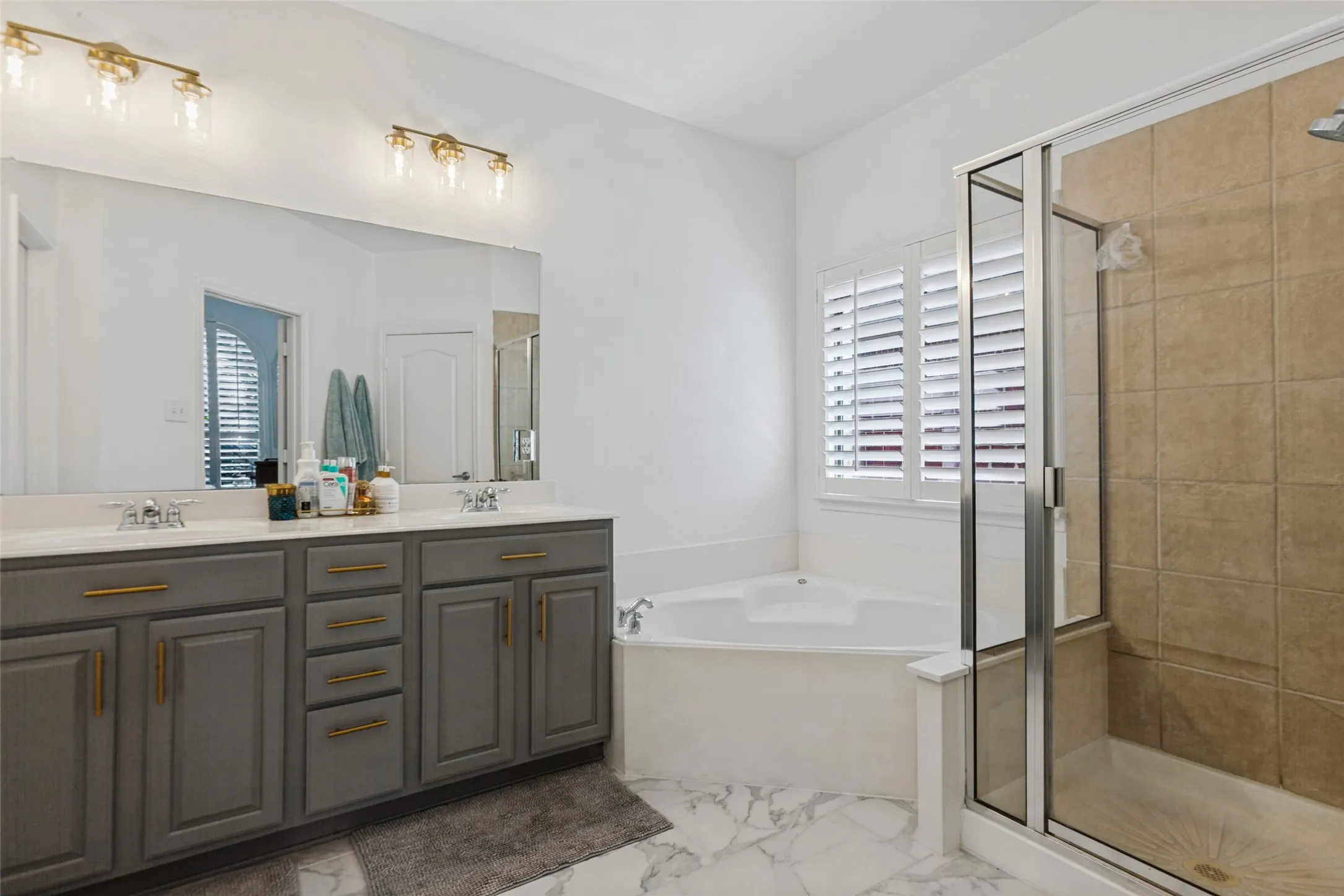 Primary ensuite bathroom has dual vanity with gray cabinetry and gold-tone hardware, large wall mirror, recessed corner bathtub, and walk-in shower with tile surround. newly tiled bathroom floor