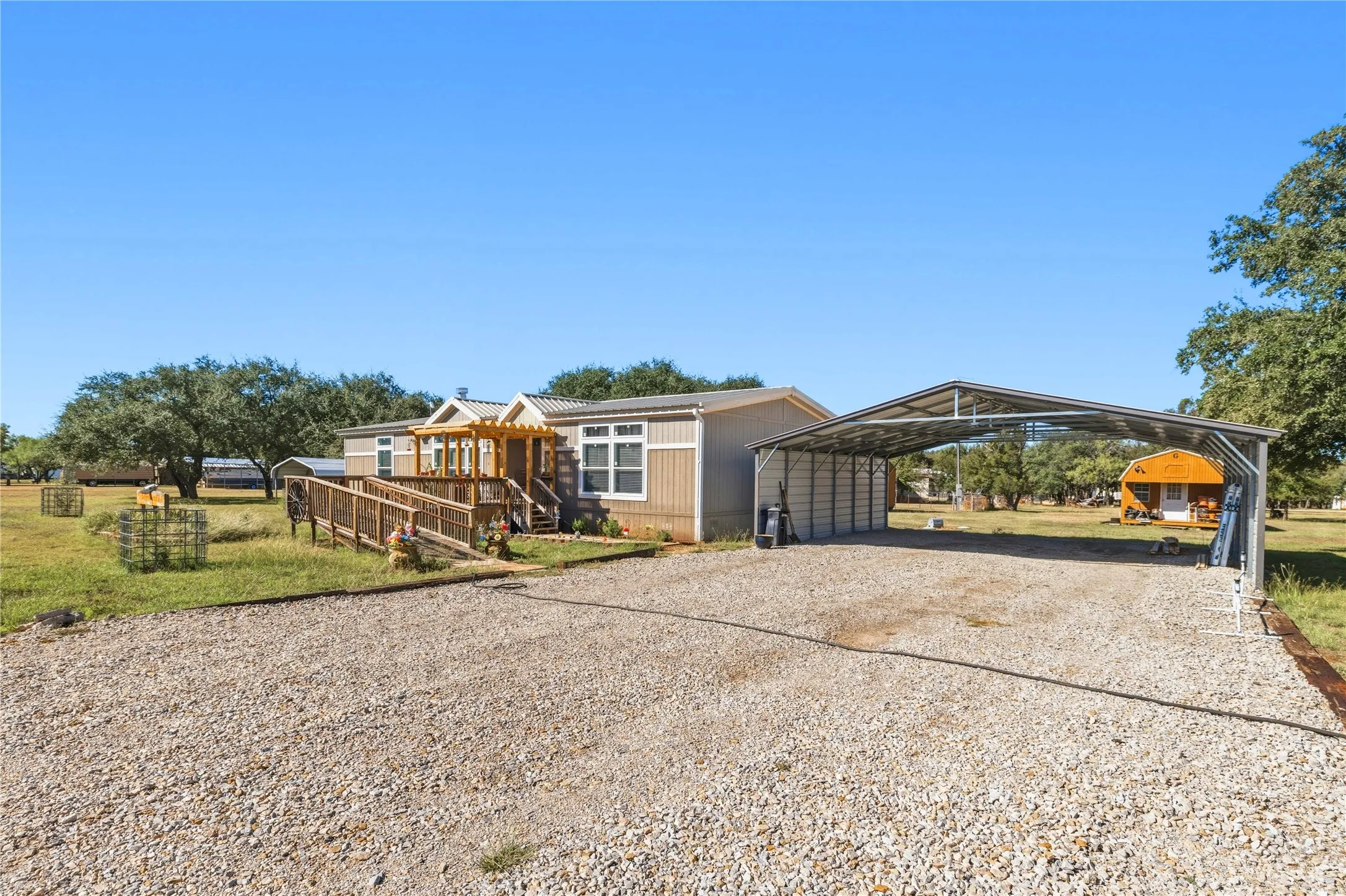 Gravel driveway leading to a metal carport, a main residence with a wooden deck and ramp, and an additional outbuilding