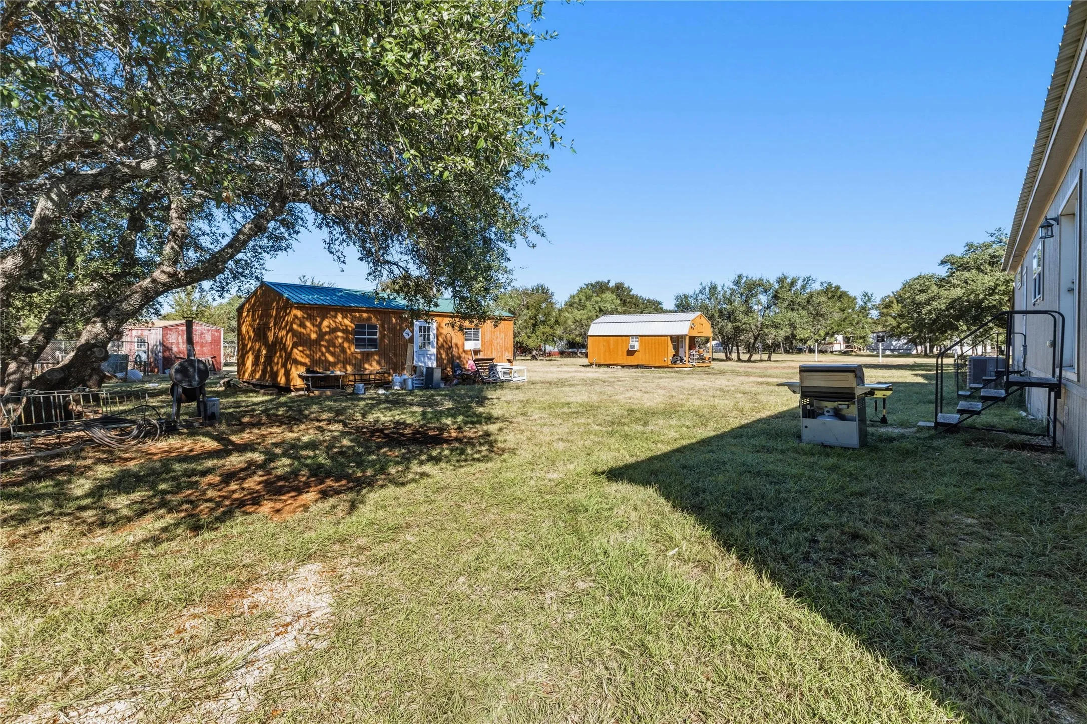 Expansive grassy lot featuring mature trees, a detached storage building with a blue roof, a second detached shed, and exterior steps leading to the main structure