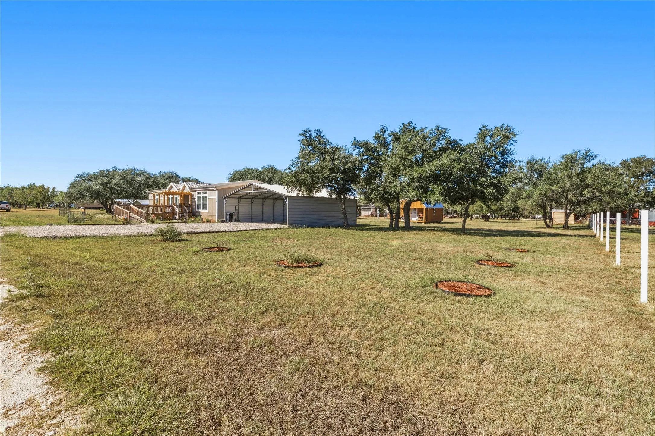 Expansive grass lot featuring mature trees, a covered carport, and a raised wooden deck with ramp access