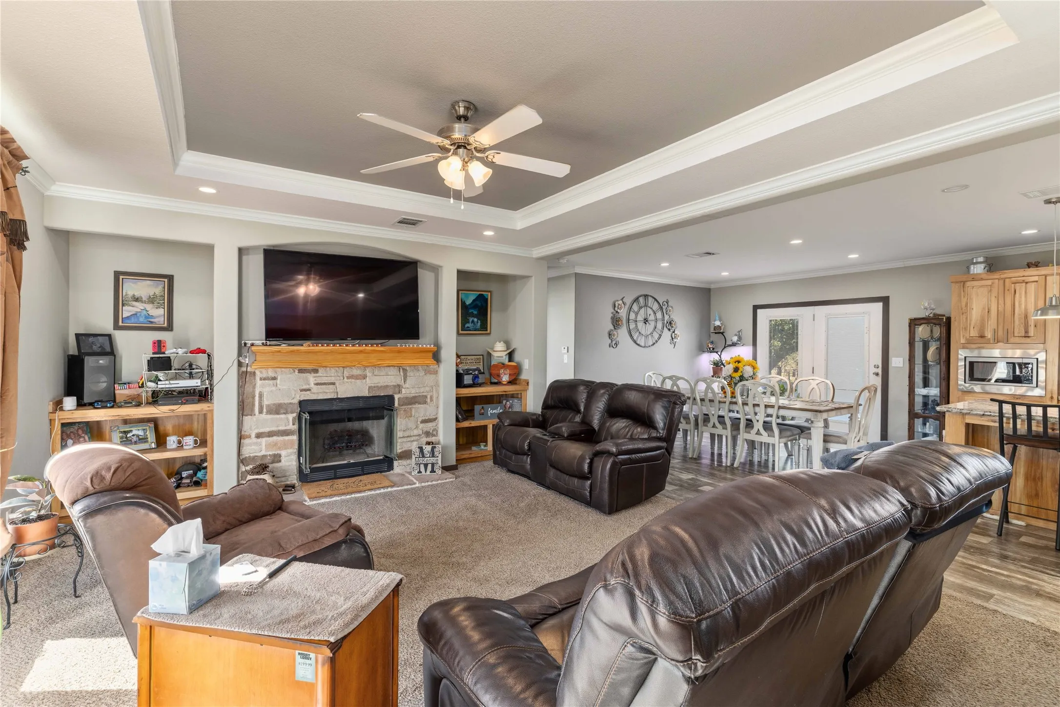 Open-concept living area featuring a stone-faced fireplace, tray ceiling with recessed lighting, and a ceiling fan