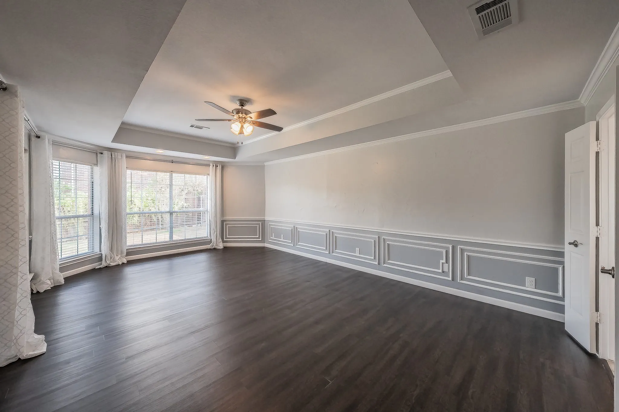 Wood-finish flooring, accent wall paneling, multi-pane windows, tray ceiling, and a ceiling fan with integrated lighting