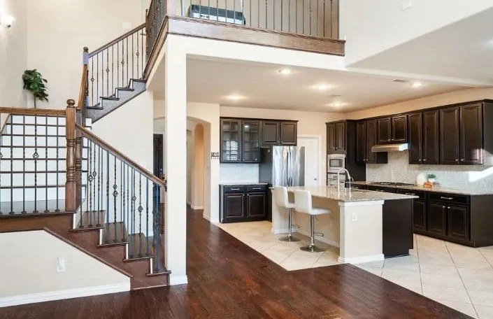 Hardwood flooring on the main level, staircase with wrought iron balusters, recessed lighting, kitchen with dark wood-finish cabinetry, and a center island with bar seating