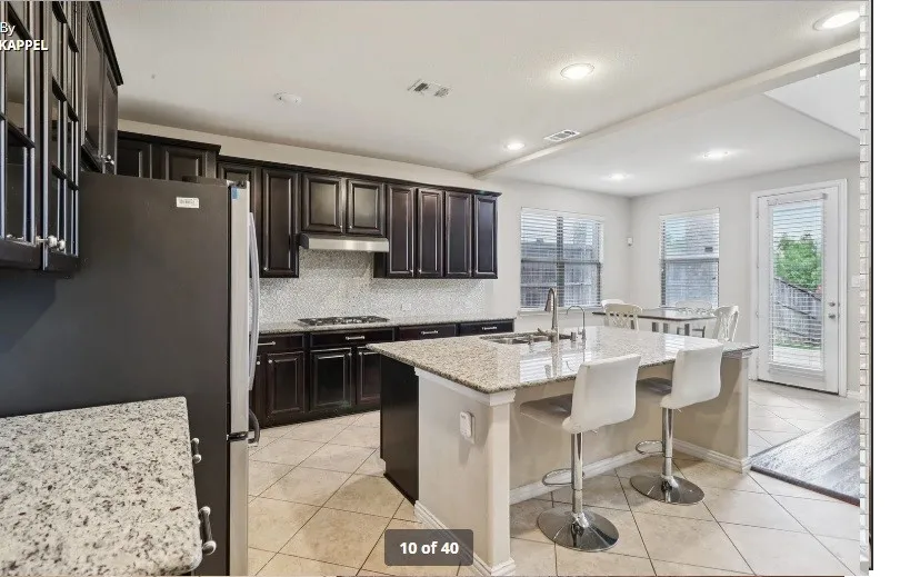Kitchen featuring dark wood cabinetry, light-toned countertops, a full backsplash, a center island with a sink, and tile flooring
