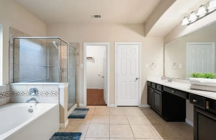 Tiled flooring, built-in soaking tub, glass-enclosed shower, dual vanity with undermount sinks, and an expansive wall mirror