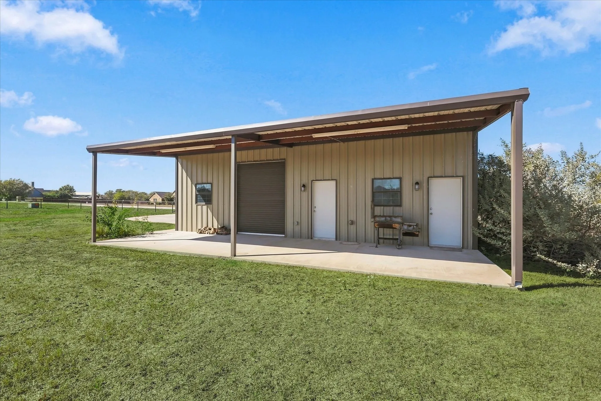Metal-sided structure featuring a covered concrete patio, an overhead garage door, two entry doors, and an exterior window