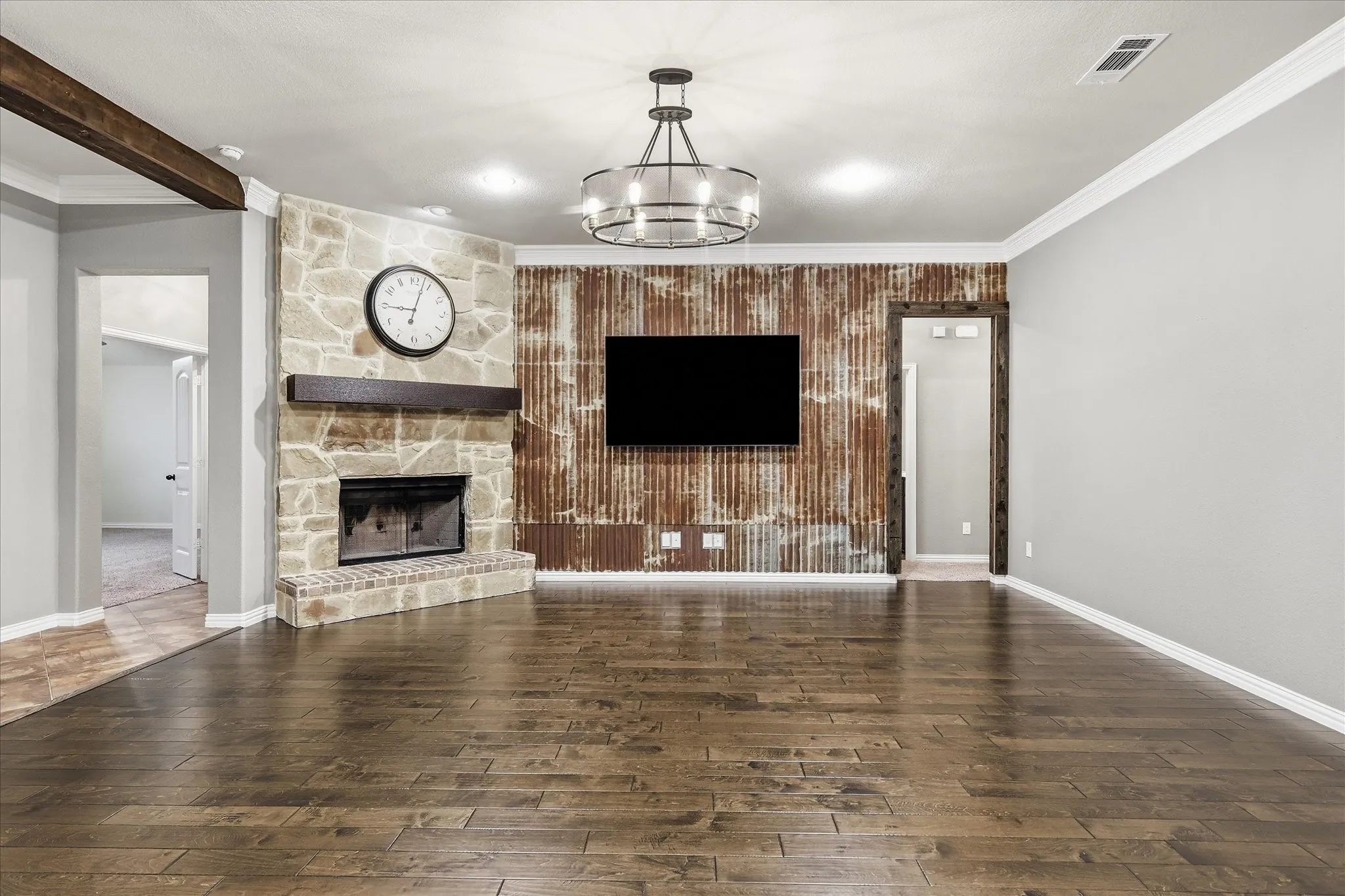 Hardwood-style flooring, stone-faced fireplace with a wood mantel, accent wall with corrugated metal paneling, wood beam ceiling accent, and a chandelier with a metal frame