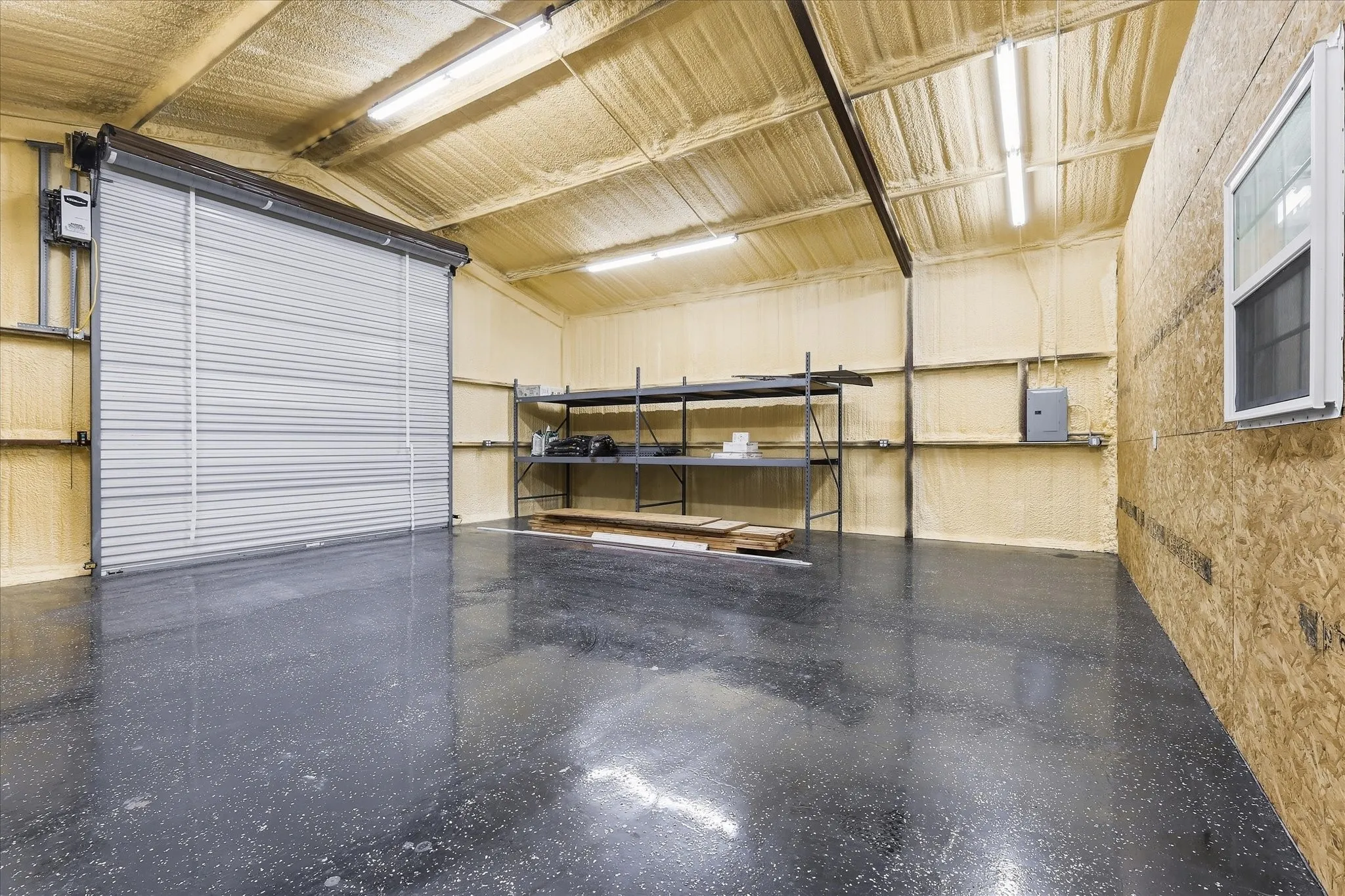 Spacious interior featuring a large roll-up door, polished concrete flooring, exposed structural beams, spray foam insulation, and a fixed window