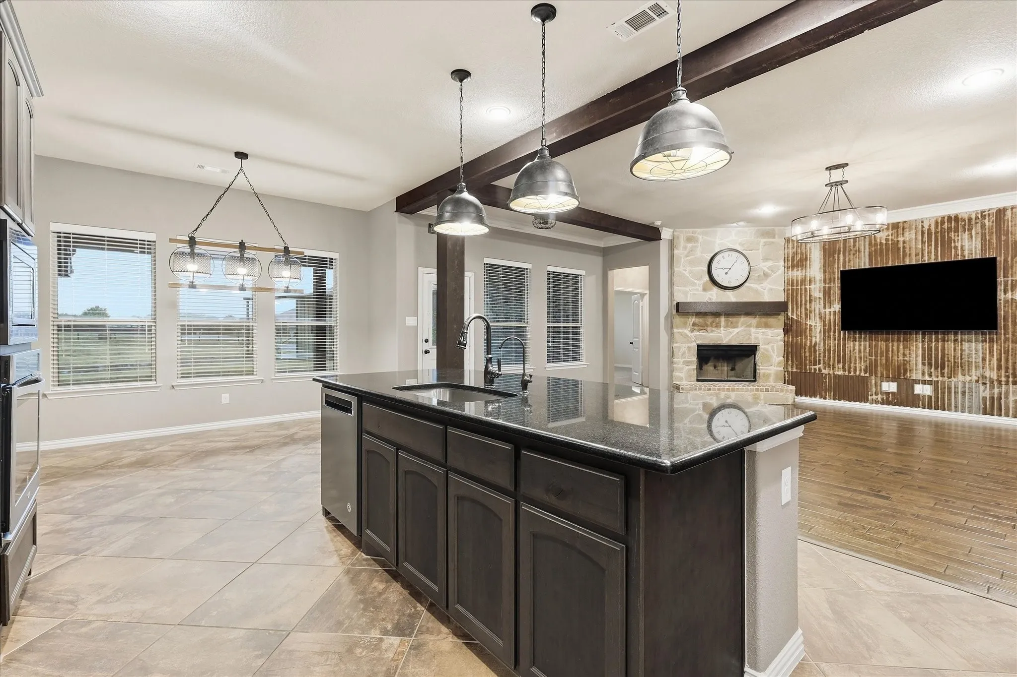 Kitchen island with dark cabinetry and a polished countertop, stainless steel dishwasher, wood-finish flooring, exposed ceiling beams, and a stone-clad fireplace