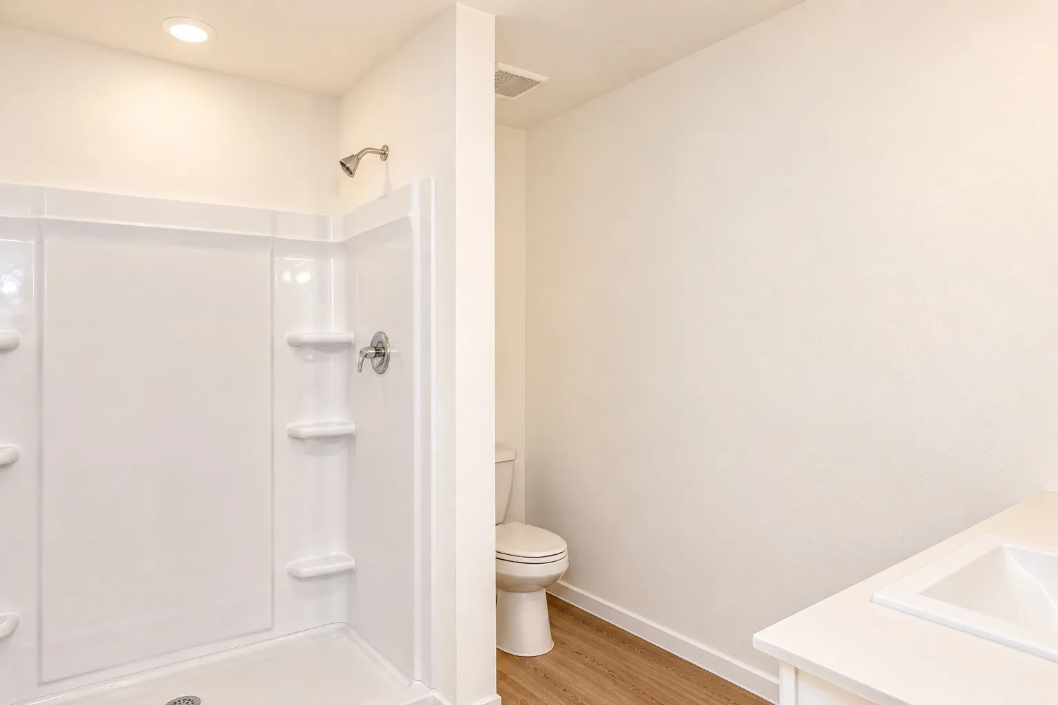 White single-piece shower surround, stainless steel shower fixtures, wood-finish flooring, white toilet, white vanity with integrated sink