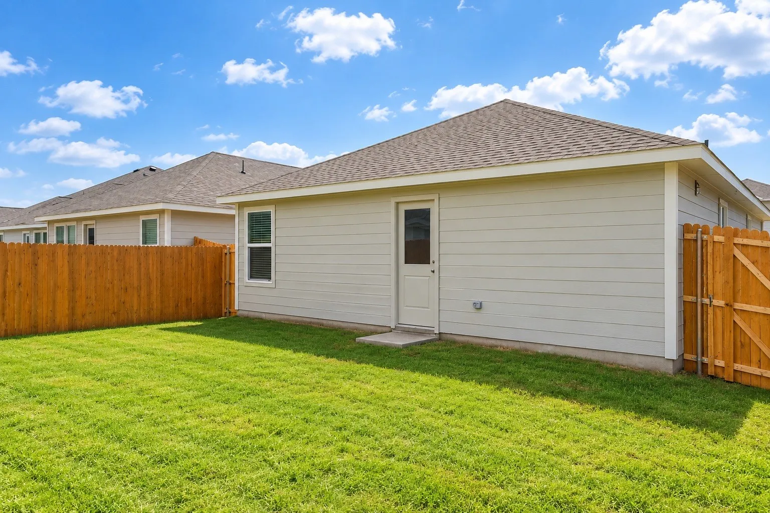 Expansive green lawn, wood fence, light-colored siding, single exterior door, and a shingle roof