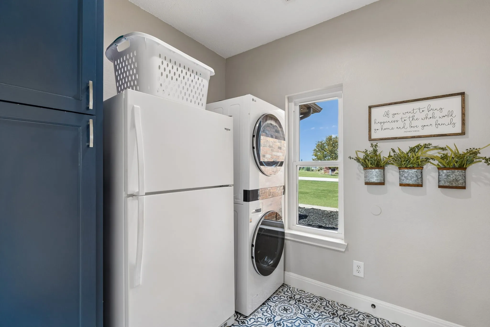 Window with exterior views, white stacked washer and dryer, patterned floor tile, and blue cabinetry