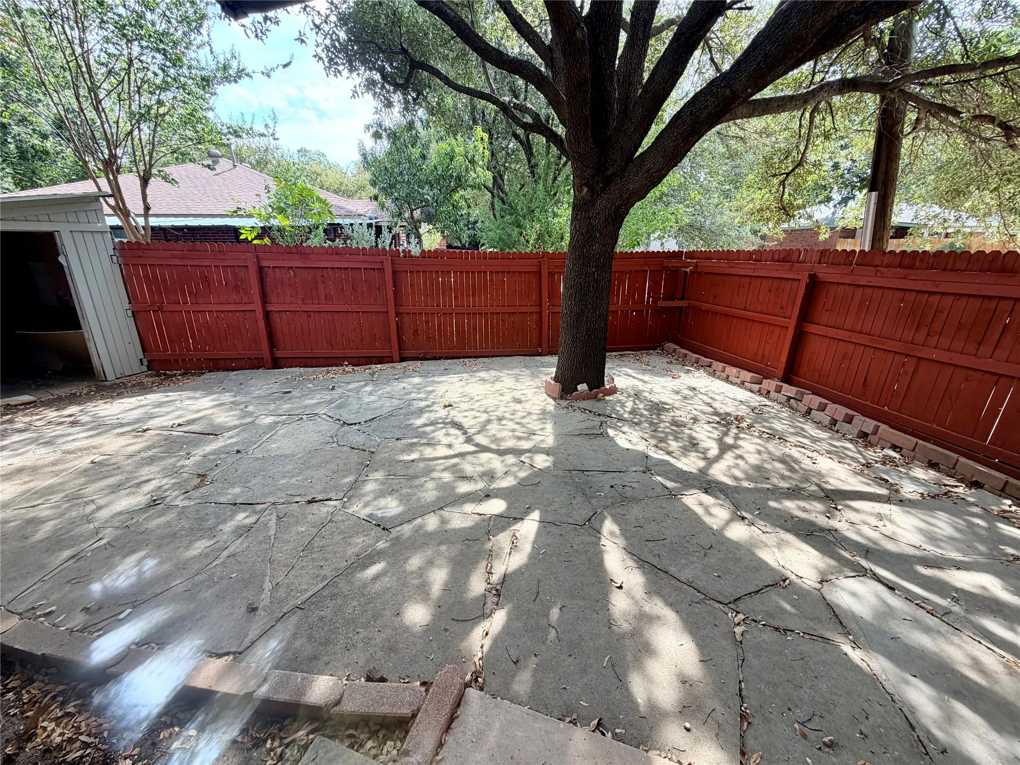 Enclosed outdoor space featuring a mature tree, red-stained wooden fence, concrete patio, and brick border