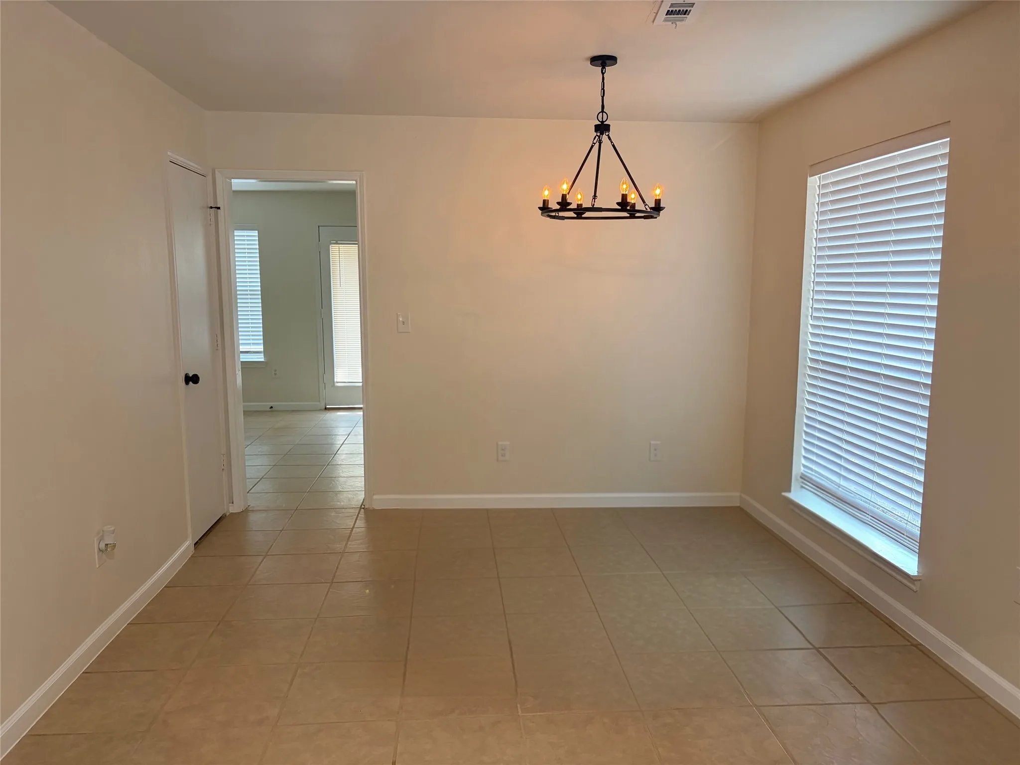 Neutral-toned walls with a black iron chandelier, tile flooring, and a large window with blinds