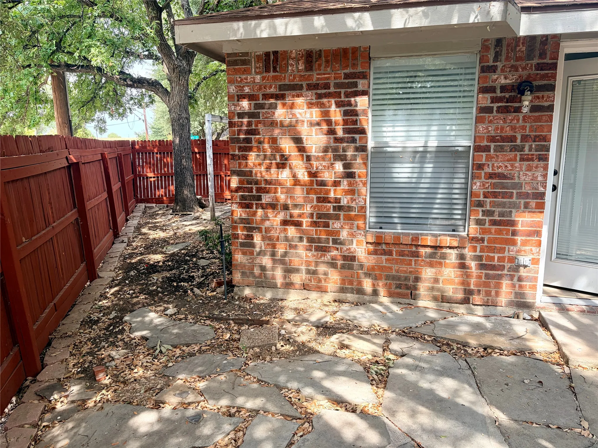 Brick exterior featuring a window with blinds, an exterior door with glass paneling, a stone patio, and a red wood fence