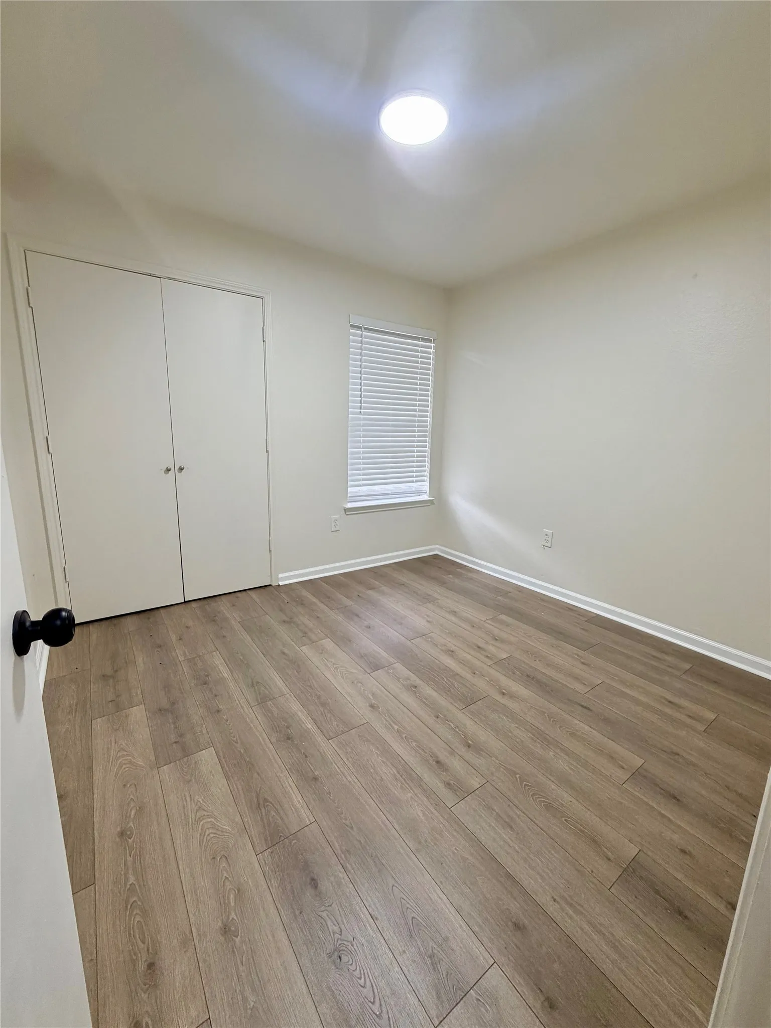 Wood-finish flooring, built-in closet with bifold doors, window with horizontal blinds, and a flush-mount ceiling light fixture