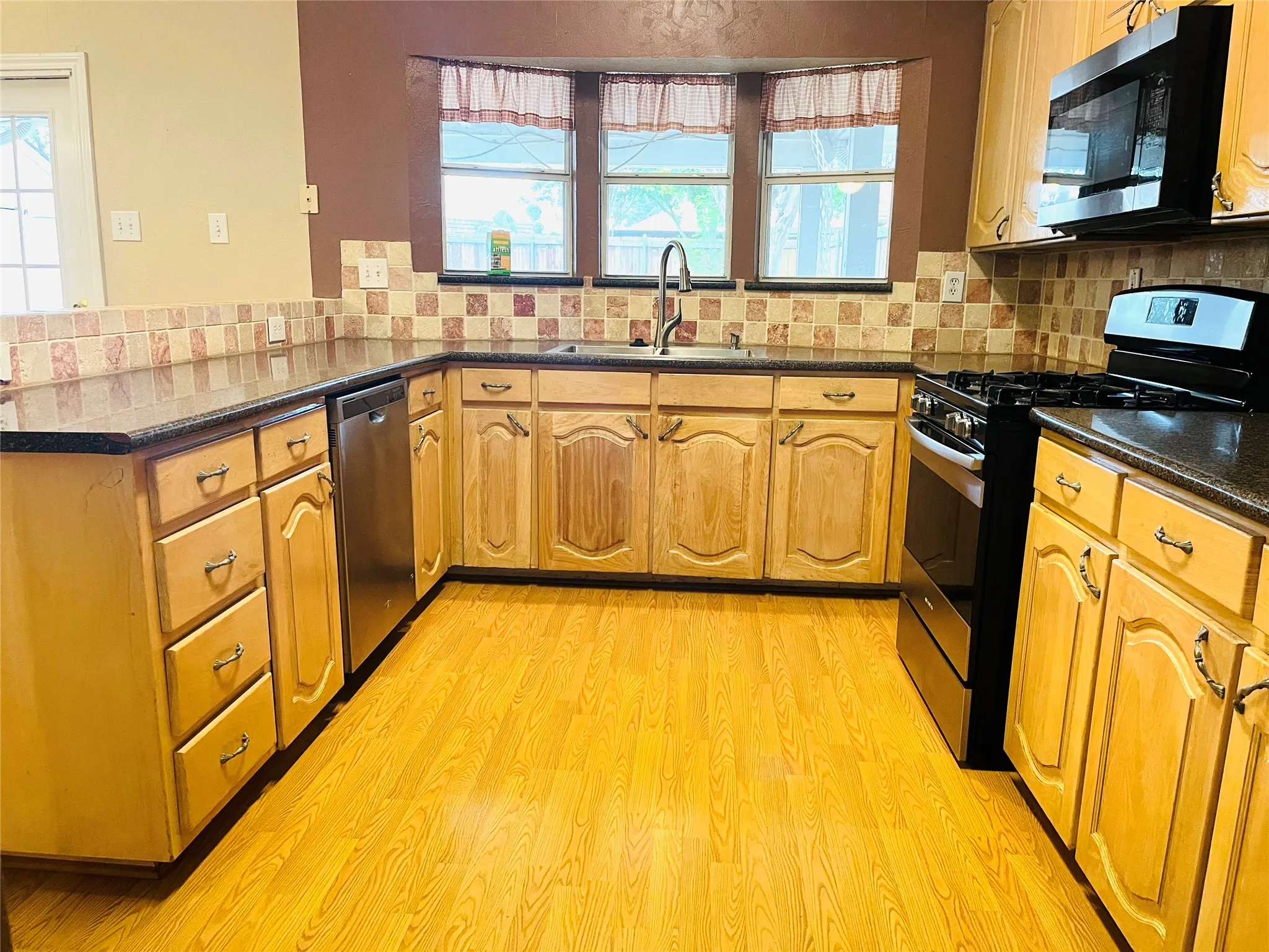 U-shaped kitchen featuring light wood cabinetry, dark countertops, tiled backsplash, and stainless steel appliances