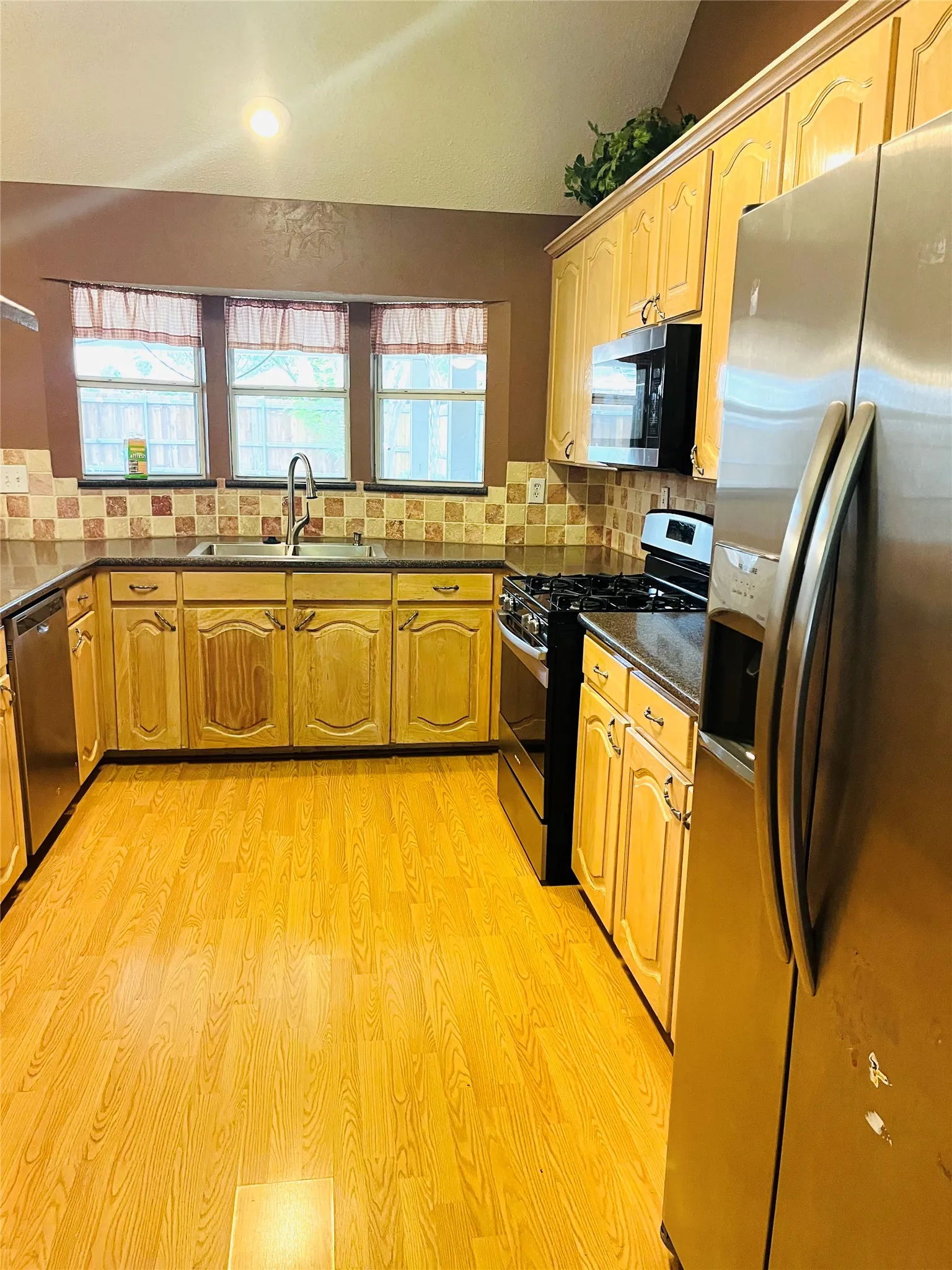 Wood-finish flooring, light wood cabinetry, stainless steel appliances, dark countertops, and a tiled backsplash