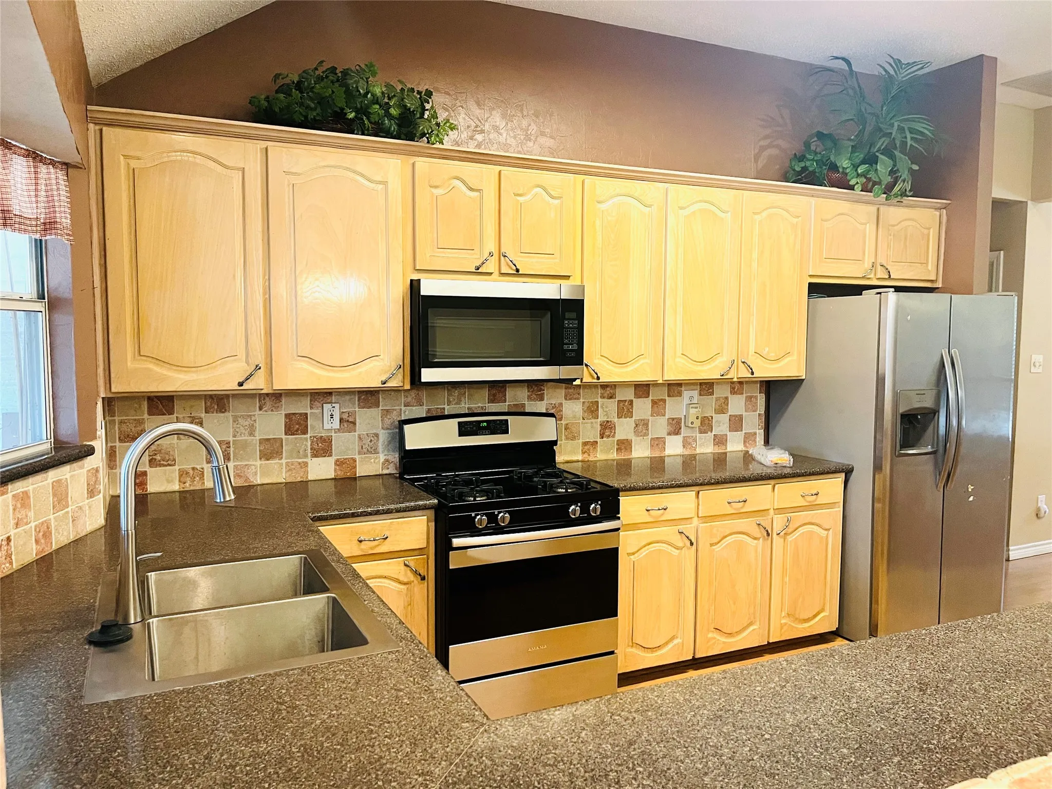 Kitchen featuring light wood-finish cabinetry, patterned tile backsplash, stainless steel gas range, built-in microwave, and a double basin stainless steel sink