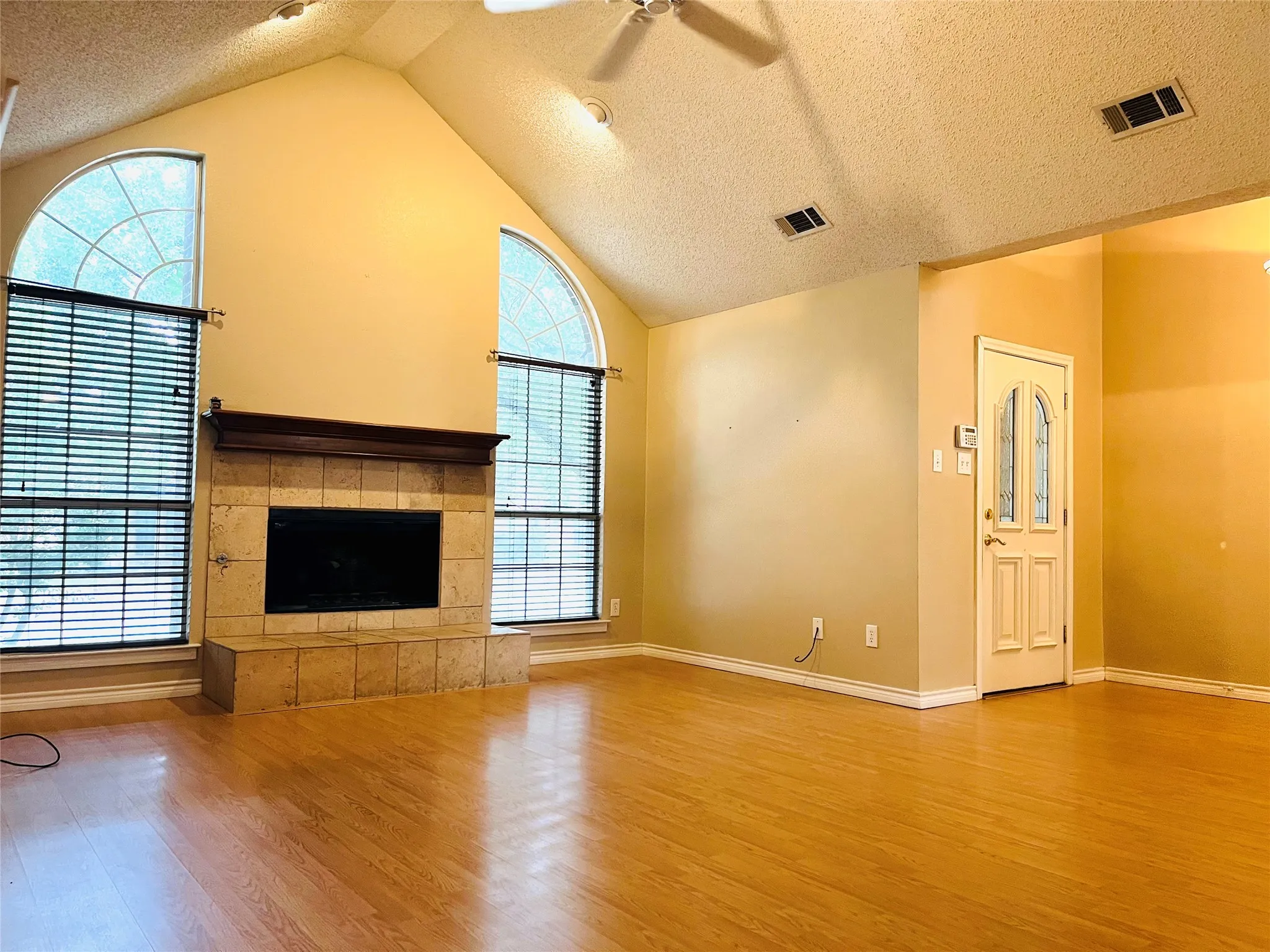 Vaulted ceiling with track lighting, wood-finish flooring, fireplace with a tiled surround, arched windows, and a decorative front door