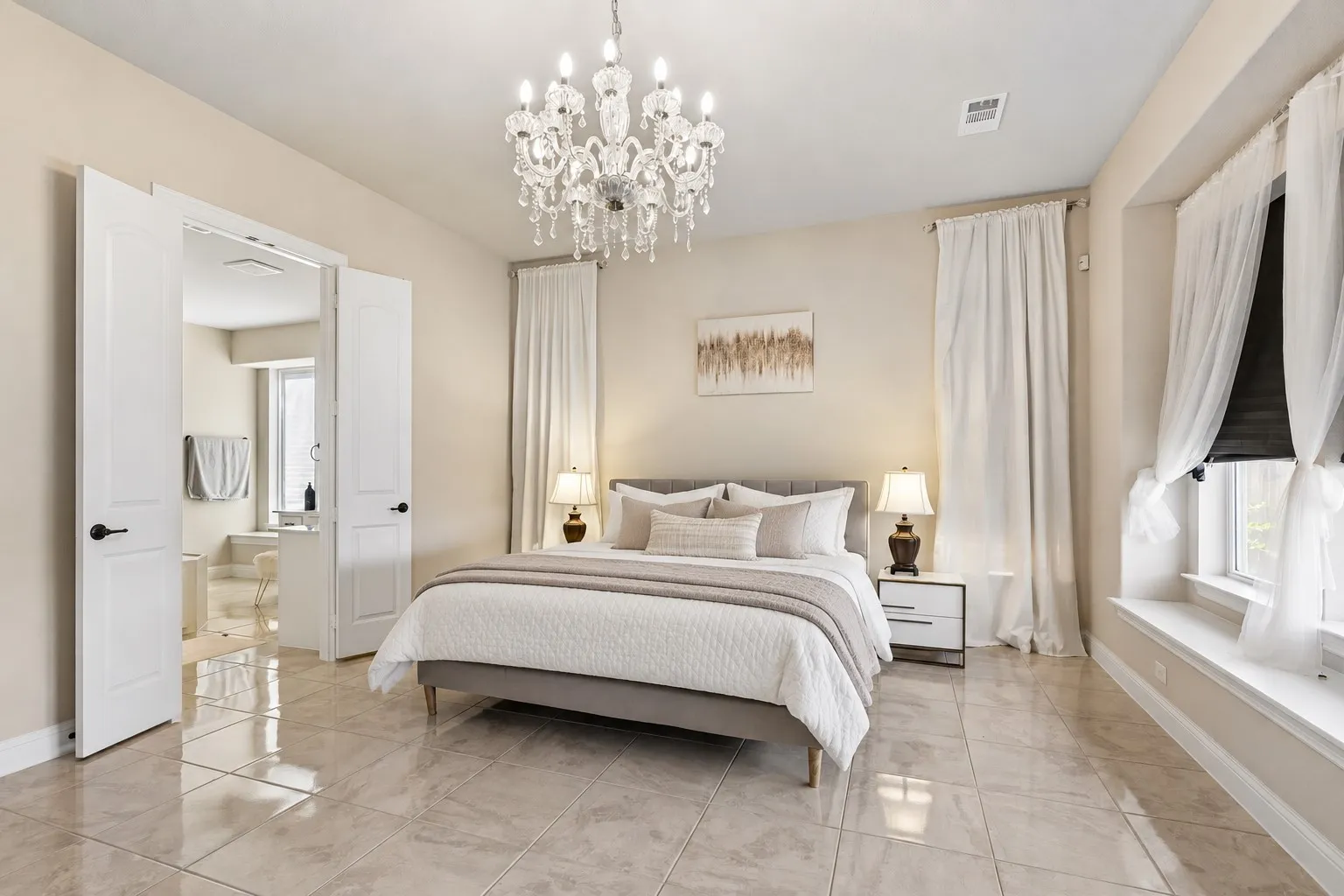 Elegant primary bedroom suite featuring soaring ceilings with crystal chandelier, tile flooring throughout, dual ensuite access via French doors, floor-to-ceiling windows with custom drapery, paired nightstands with integrated lighting, and a spacious layout accommodating a substantial bed frame