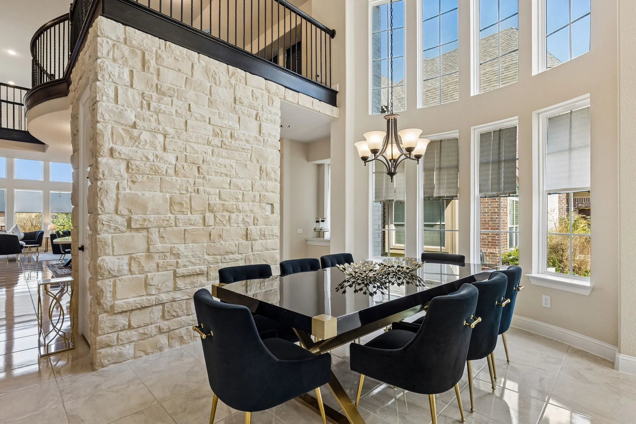Two-story stone accent wall, upper-level balcony with dark wood railing, two-story window array, tile flooring, and an ornate chandelier