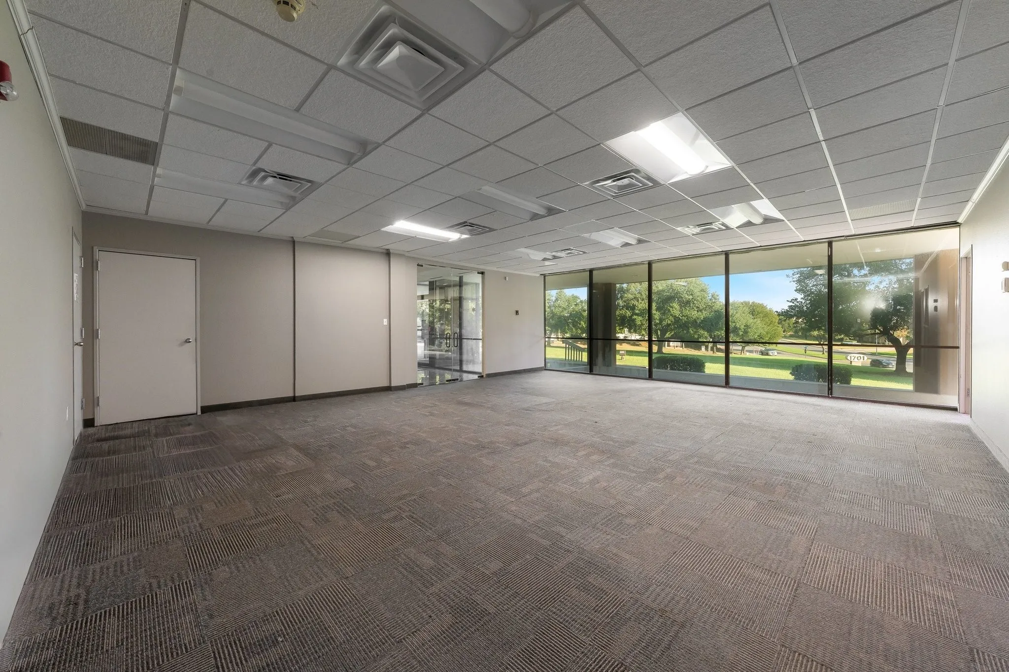 Expansive commercial space featuring wall-to-wall carpeting, a grid-pattern drop ceiling, recessed lighting, and large windows with exterior green space