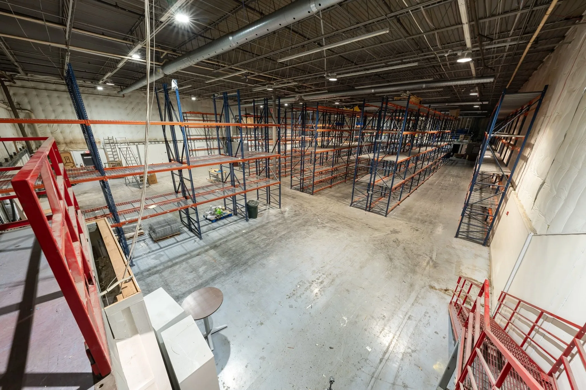 Warehouse space featuring exposed ceiling infrastructure, overhead lighting, concrete flooring, extensive pallet racking, and a red metal staircase